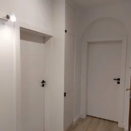 Wnętrze przedpokoju z dwiema białymi, minimalistycznymi drzwiami z czarnymi klamkami oraz białą szafą w zabudowie, oświetlone punktowym światłem.