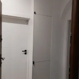 Wysoka, zabudowana szafa wnękowa z trzema segmentami, w kolorze jasnoszarym, z minimalistycznymi, czarnymi uchwytami, wkomponowana w korytarz z białymi ścianami i widocznymi drzwiami po lewej...