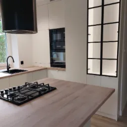 Nowoczesna, jasna kuchnia z wyspą, czarną płytą grzewczą i okapem cylindrycznym. Na ścianie przeszklona witryna z podświetleniem.