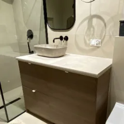 Nowoczesna łazienka z szafką pod umywalkę z drewnianymi frontami i kamiennym blatem, minimalistyczna armatura w czarnym macie, owalne lustro w czarnej ramie i geometryczna lampa wisząca z żarówką...