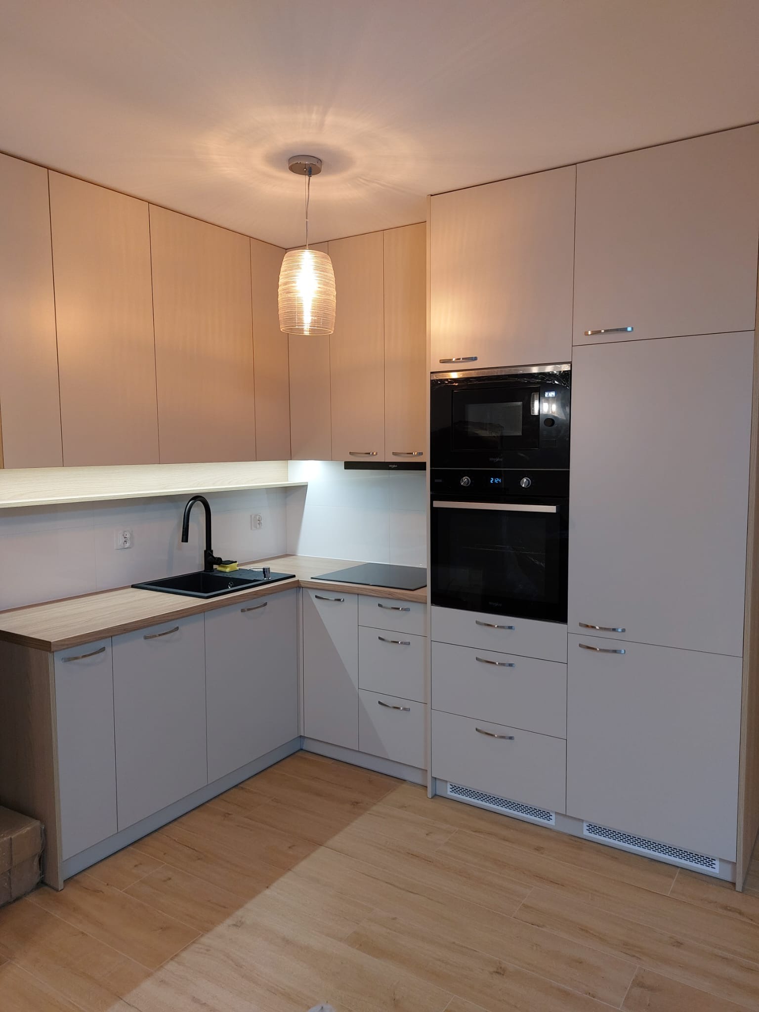 Jasna kuchnia z drewnianym blatem, czarnym zlewem i piekarnikiem w zabudowie, oświetlona wiszącą lampą z żarnika.