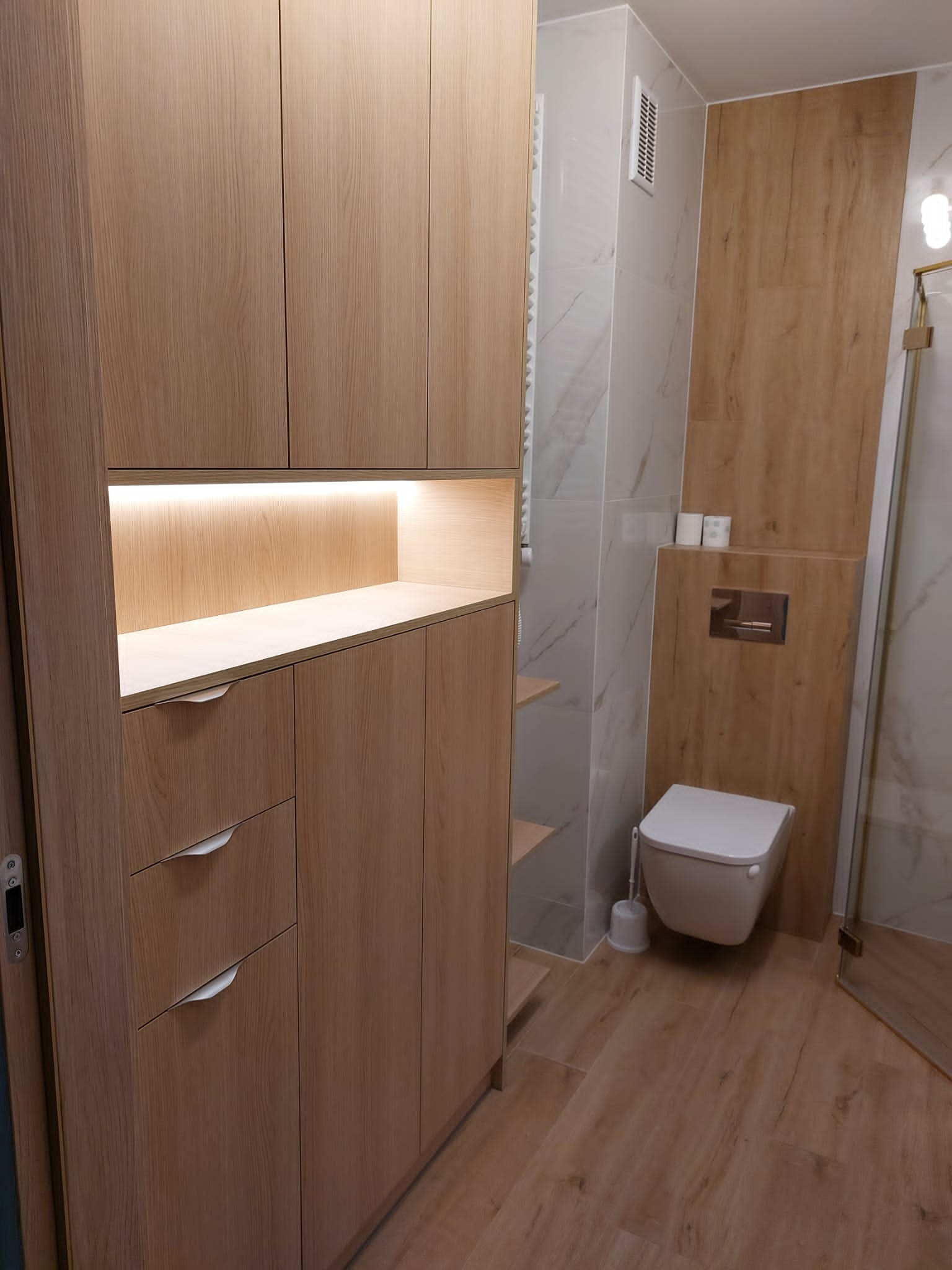 Jasna, drewniana zabudowa meblowa w łazience z podświetlaną półką, dopasowana do drewnopodobnych płytek podłogowych i ściennych, z widoczną toaletą podwieszaną i częściowo otwartą kabiną prysznicową.