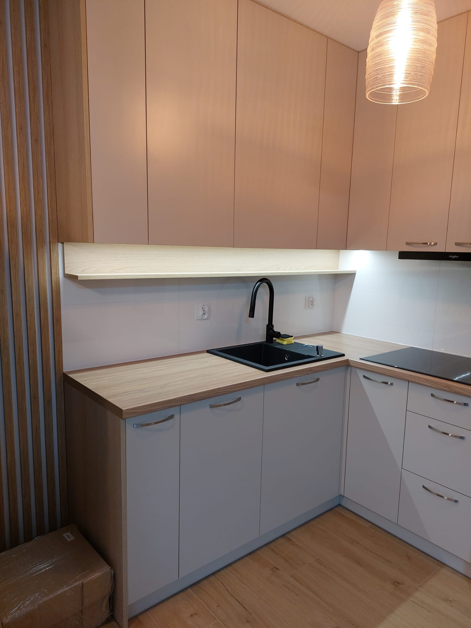 Jasna kuchnia narożna z drewnianym blatem i czarnym zlewem, oświetlona wiszącą lampą z plecionki, z dekoracyjną ścianką z drewnianych listewek po lewej stronie i kartonem na podłodze.