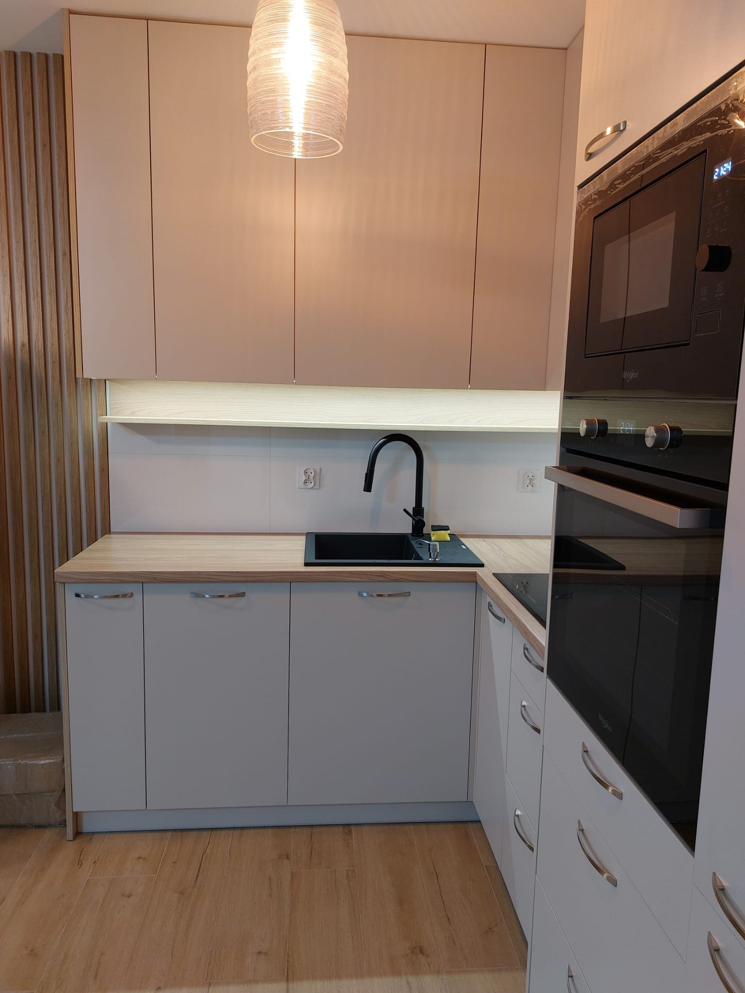 Jasna kuchnia z drewnianymi akcentami: szafki górne w odcieniach beżu, drewniany panel ścienny, czarny zlew i bateria, piekarnik w zabudowie, drewniana podłoga, oświetlenie punktowe pod szafkami.