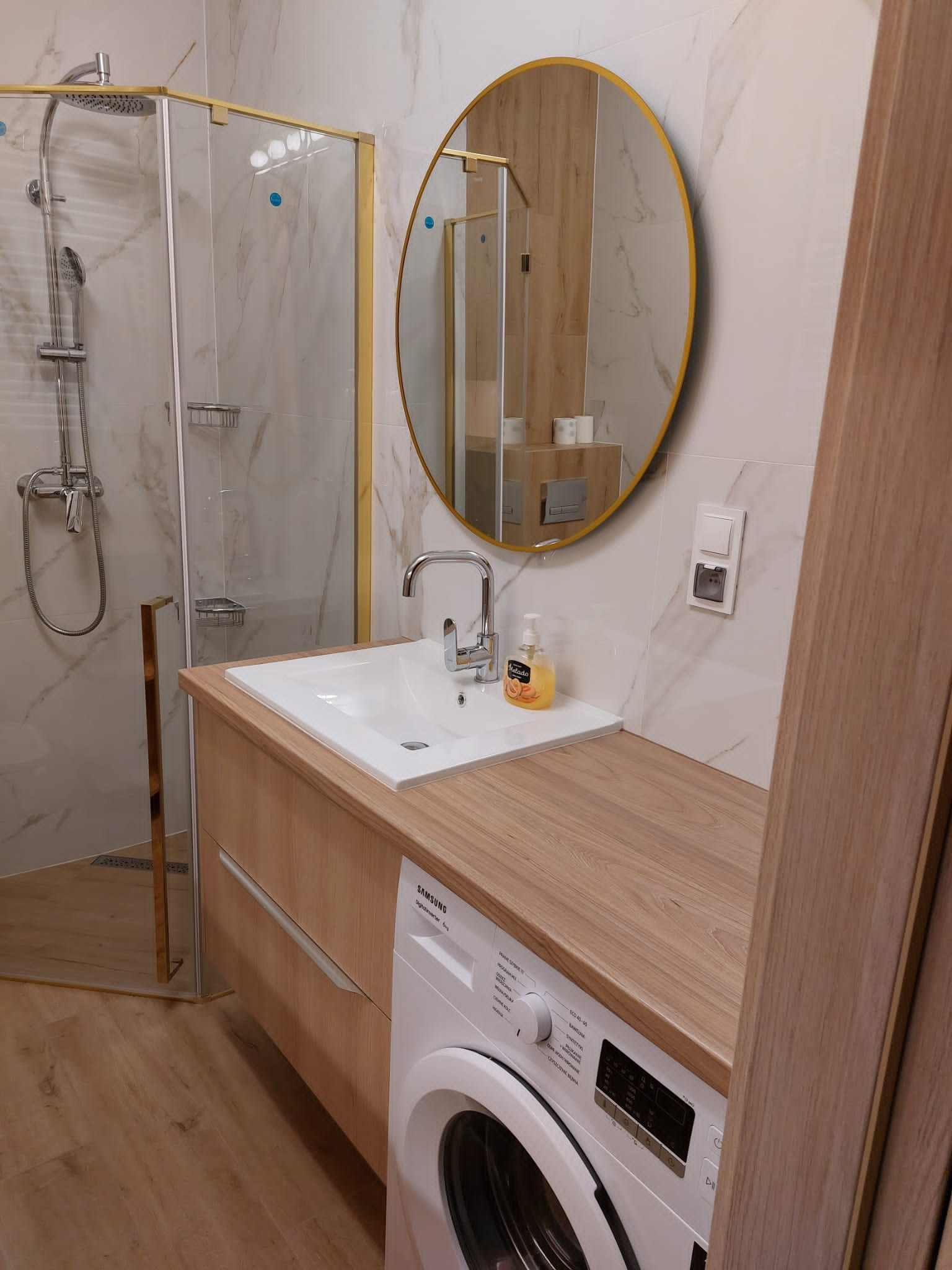 Nowoczesna łazienka z kabiną prysznicową w złotej ramie, okrągłym lustrem z ramą w kolorze złota, umywalką nablatową na szafce z blatem imitującym drewno, pralką pod blatem i marmurowymi płytkami...