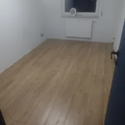 Ułożone panele podłogowe w pokoju z białymi ścianami, grzejnikiem i małym wentylatorem na parapecie okna.