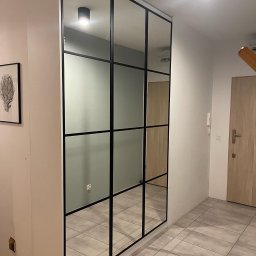 HQRZieliński Robert Zieliński - Duża lustrzana zabudowa z czarnymi szprosami w przedpokoju, odbijająca fragment wnętrza i oświetlenie sufitowe.