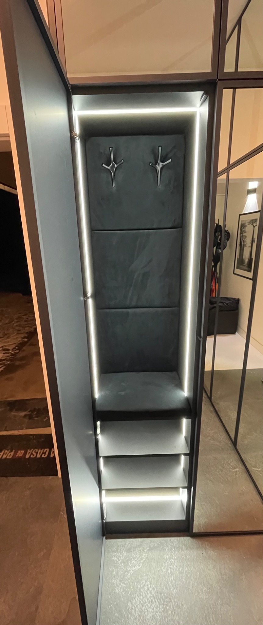 Otwarta szafa wnękowa z podświetleniem LED, wykończona szarą tapicerką i metalowymi wieszakami, widoczna na tle lustrzanych drzwi.