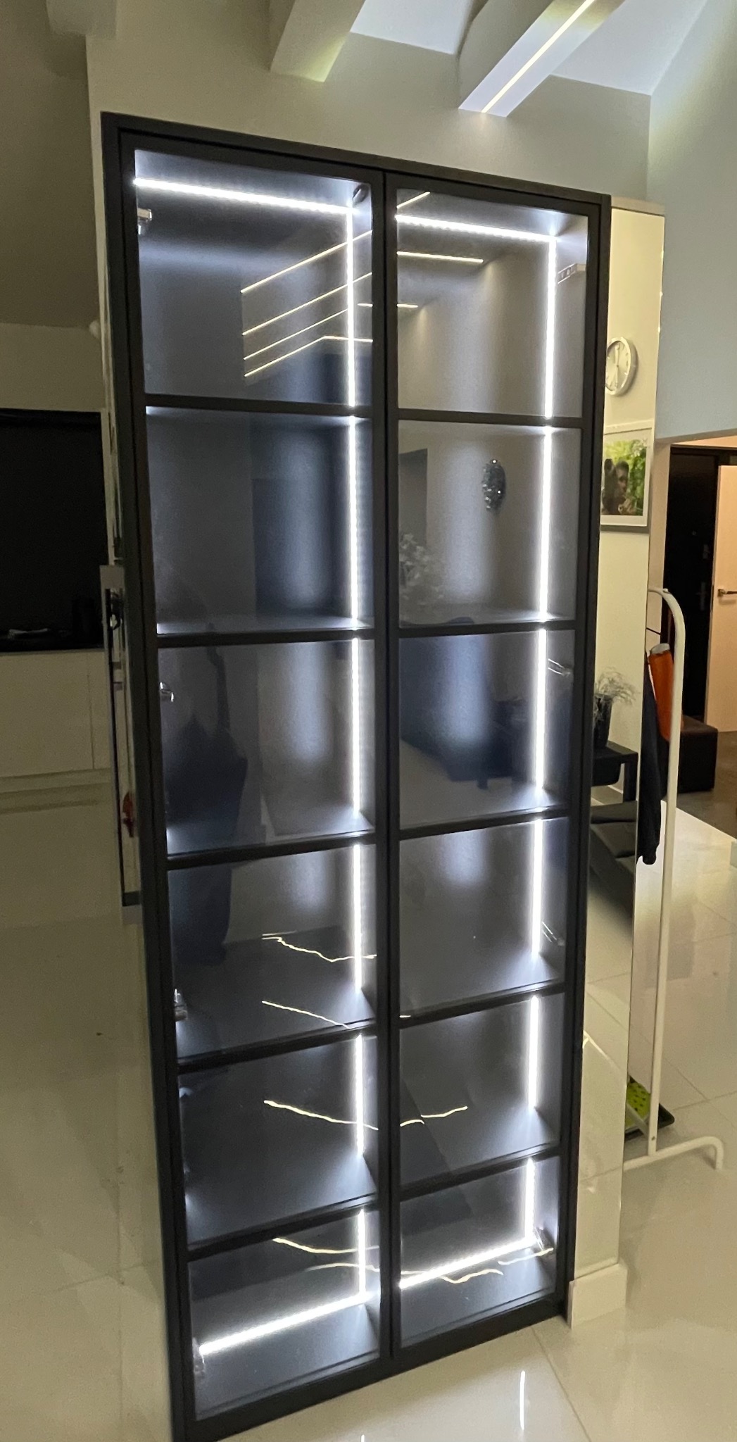 Dwudrzwiowa, wysoka witryna z czarnymi ramami i oświetleniem LED na każdej półce, w minimalistycznym wnętrzu.