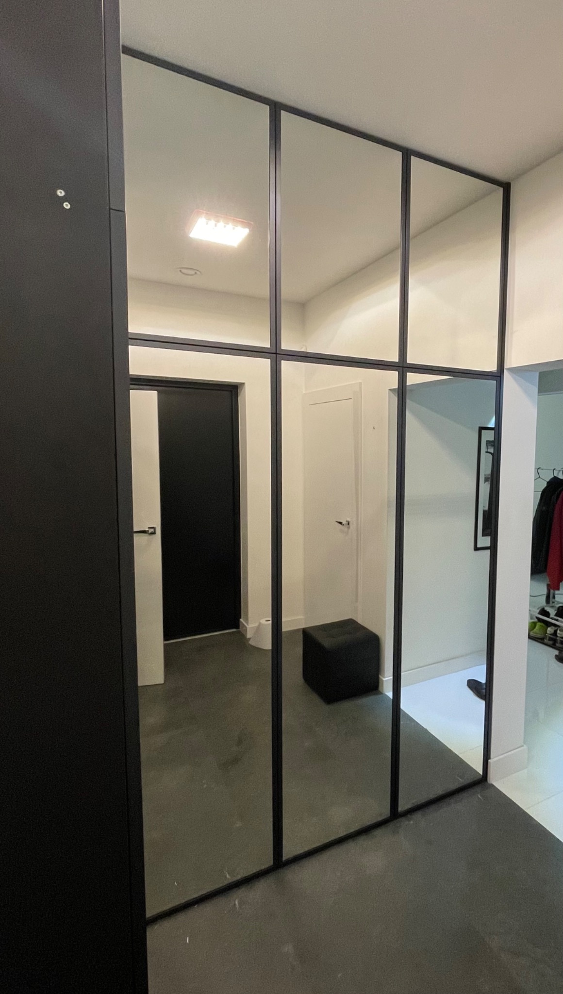 Szafa wnękowa z lustrzanymi drzwiami i czarnymi ramami w przedpokoju w Warszawie, odbijająca drzwi i pufę.