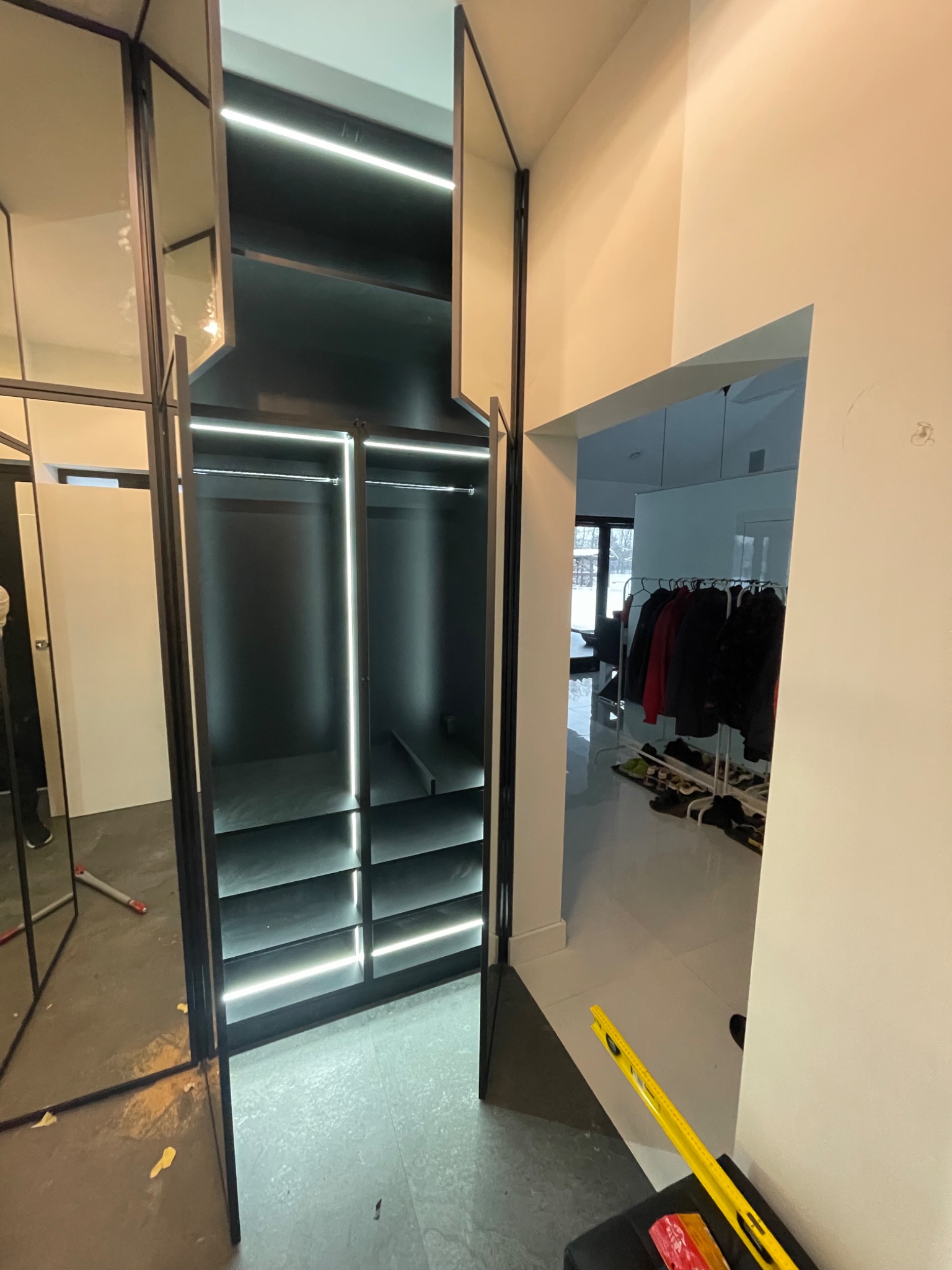 Nowoczesna, czarna garderoba z oświetleniem LED, częściowo otwarta, widok na wieszaki z ubraniami w tle.