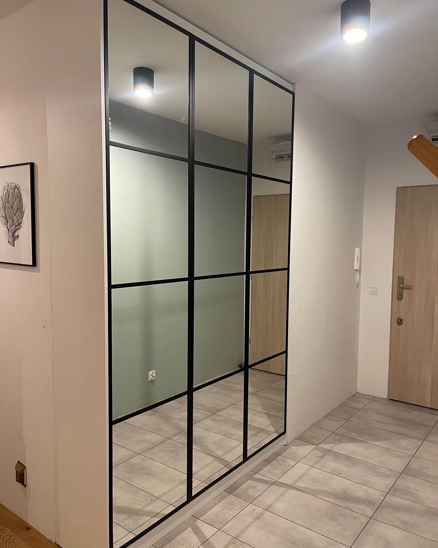 Duża lustrzana zabudowa z czarnymi szprosami w przedpokoju, odbijająca fragment wnętrza i oświetlenie sufitowe.