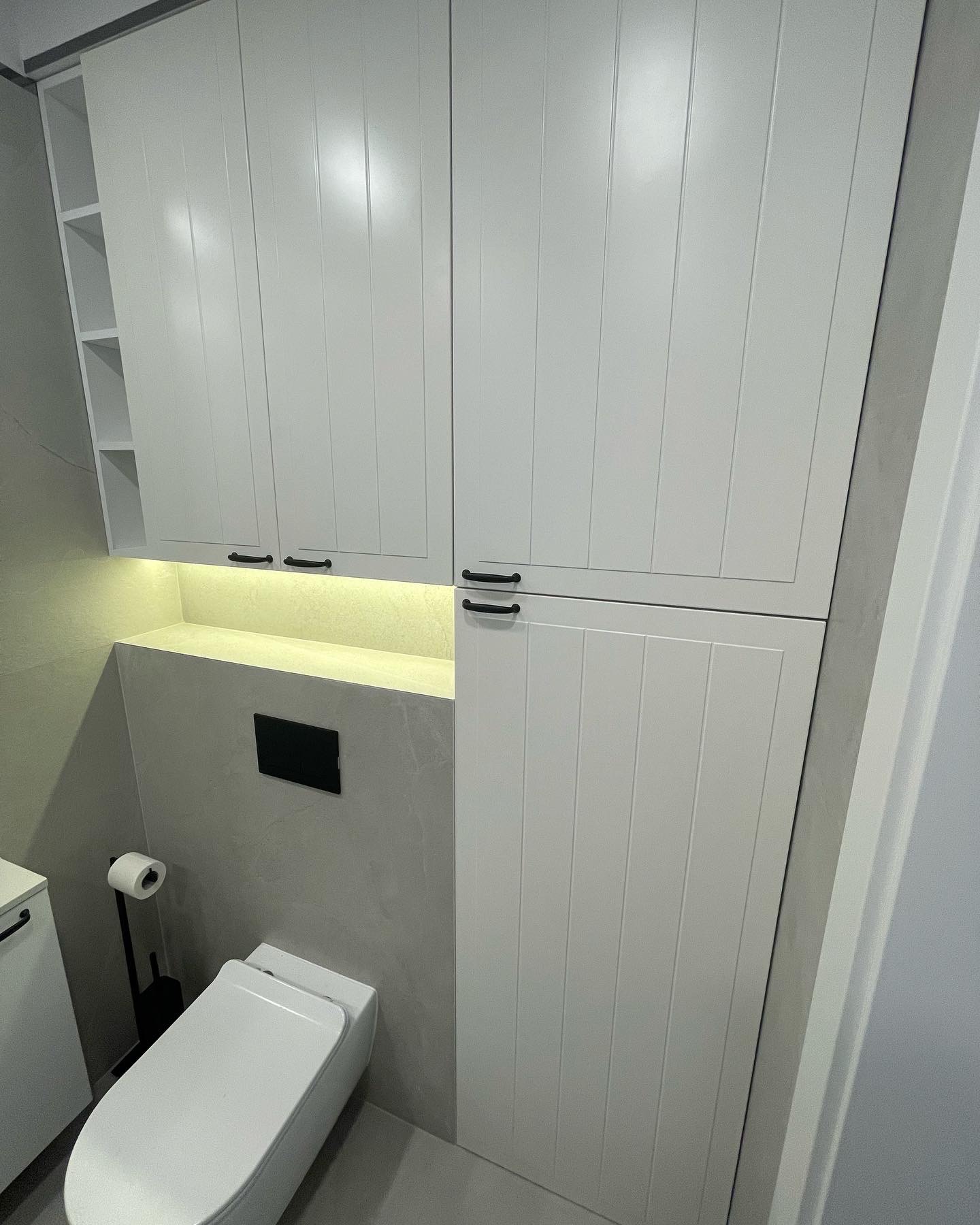 Biała zabudowa toalety z szafkami z frezowanymi frontami i czarnymi uchwytami, podświetlona półka nad stelażem WC, minimalistyczny design.