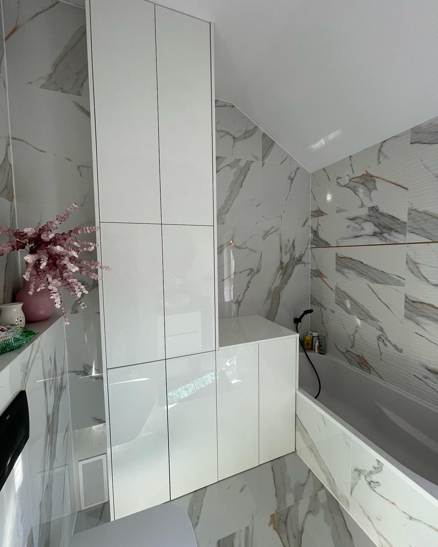 Biała, wysoka szafa łazienkowa z błyszczącymi frontami, dopasowana do skosu dachu, z dekoracją w postaci różowego wazonu z kwiatami na blacie wykończonym marmurem.