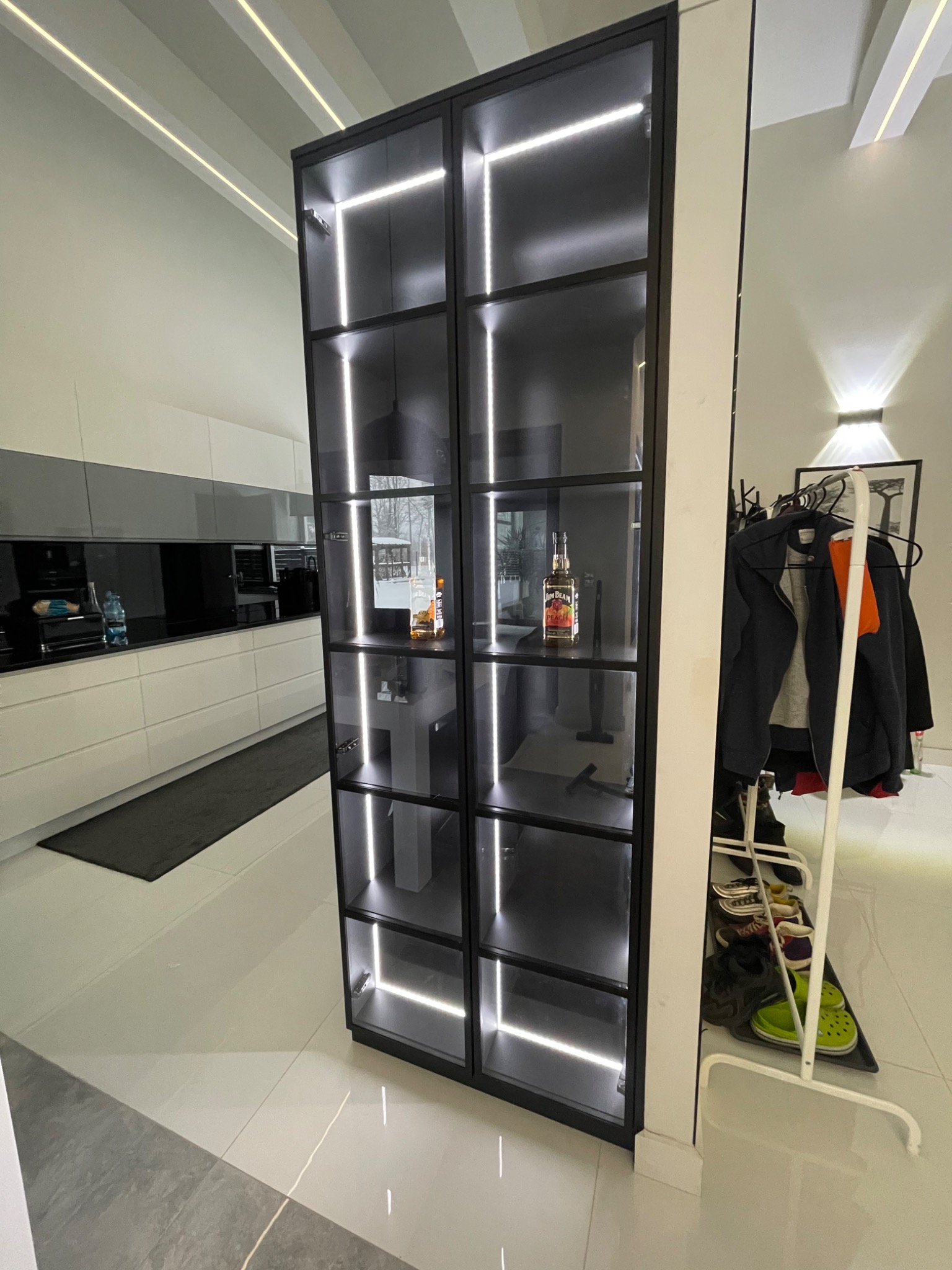 Nowoczesna, czarna witryna z podświetlanymi półkami, zintegrowana z zabudową kuchenną, widoczna butelka alkoholu na jednej z półek, obok wieszak na ubrania.