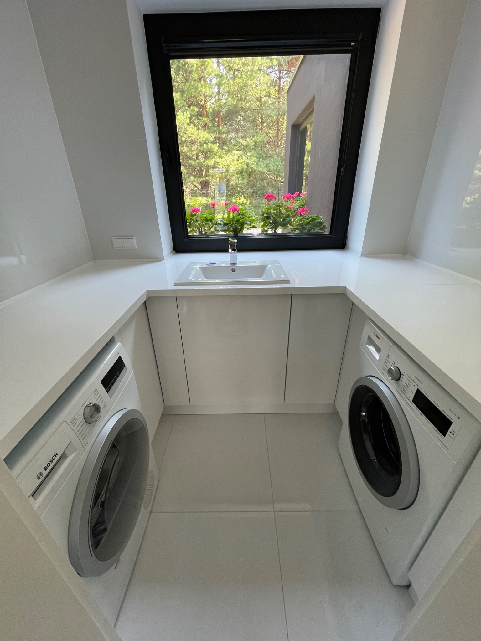 Biała, nowoczesna pralnia w skosie dachu z dwoma pralkami Bosch po bokach, blatem z umywalką pod oknem z widokiem na zieleń i różowe kwiaty.