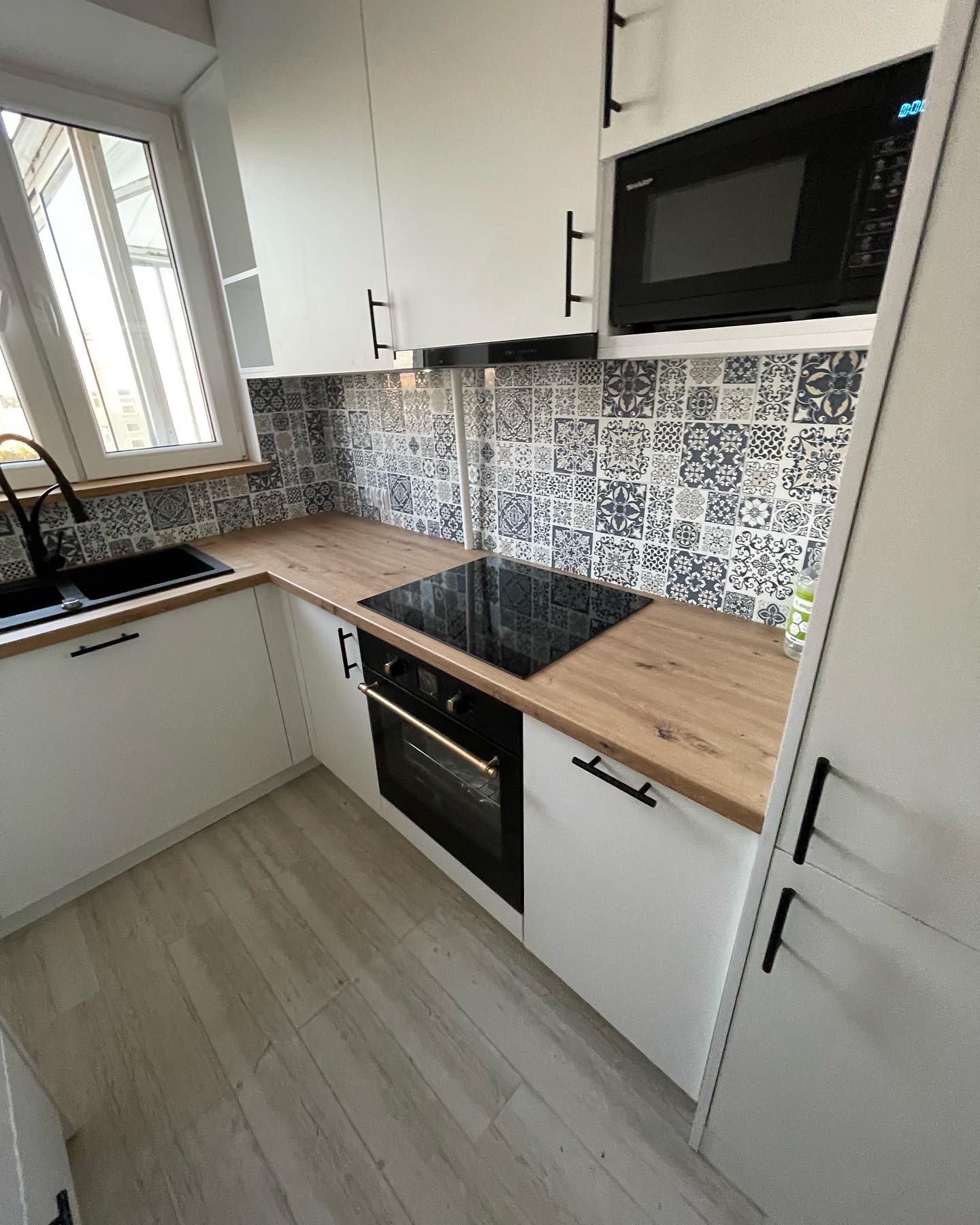 Nowoczesna, jasna kuchnia z białymi szafkami, drewnianym blatem i czarną armaturą, płyta indukcyjna i dekoracyjne płytki nad blatem.