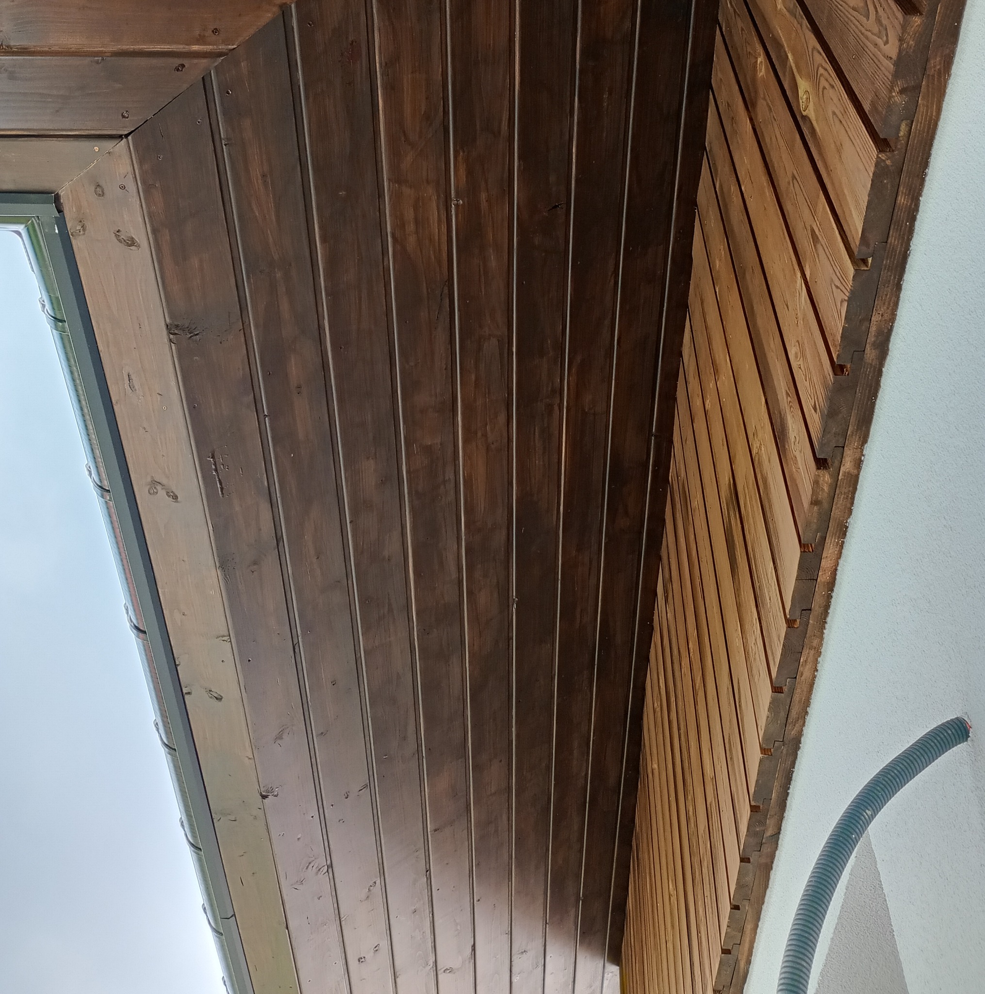 Drewniana podbitka dachowa, pomalowana na ciemny brąz, widoczna od dołu pod kątem. Fragment rynny i ściany budynku w tle. Widoczne łączenia desek i struktura drewna.