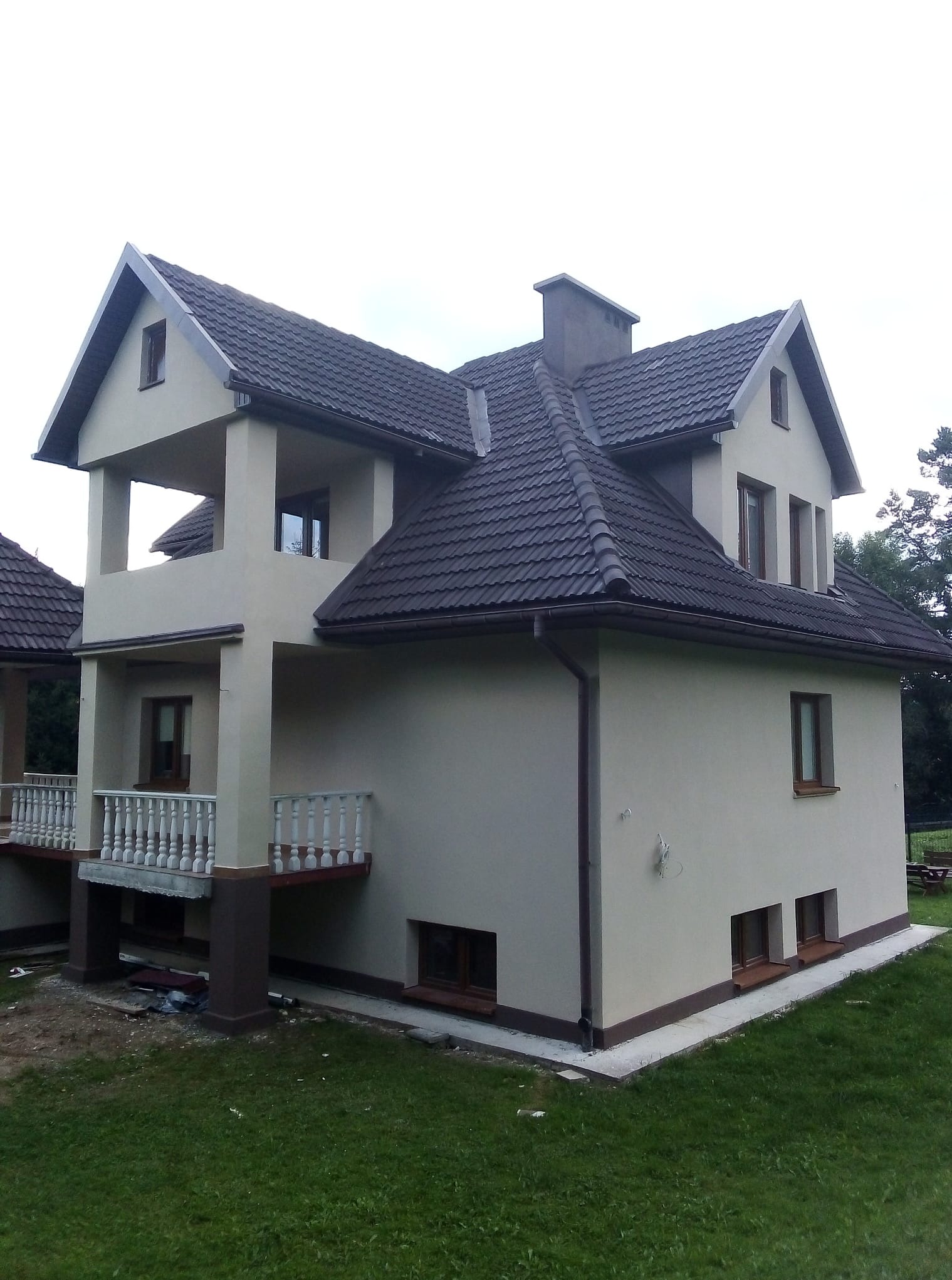 Dwukondygnacyjny dom z beżową elewacją, ciemnobrązowym dachem i balkonem z białą balustradą. Widok z boku na tle zielonego trawnika i drzew.