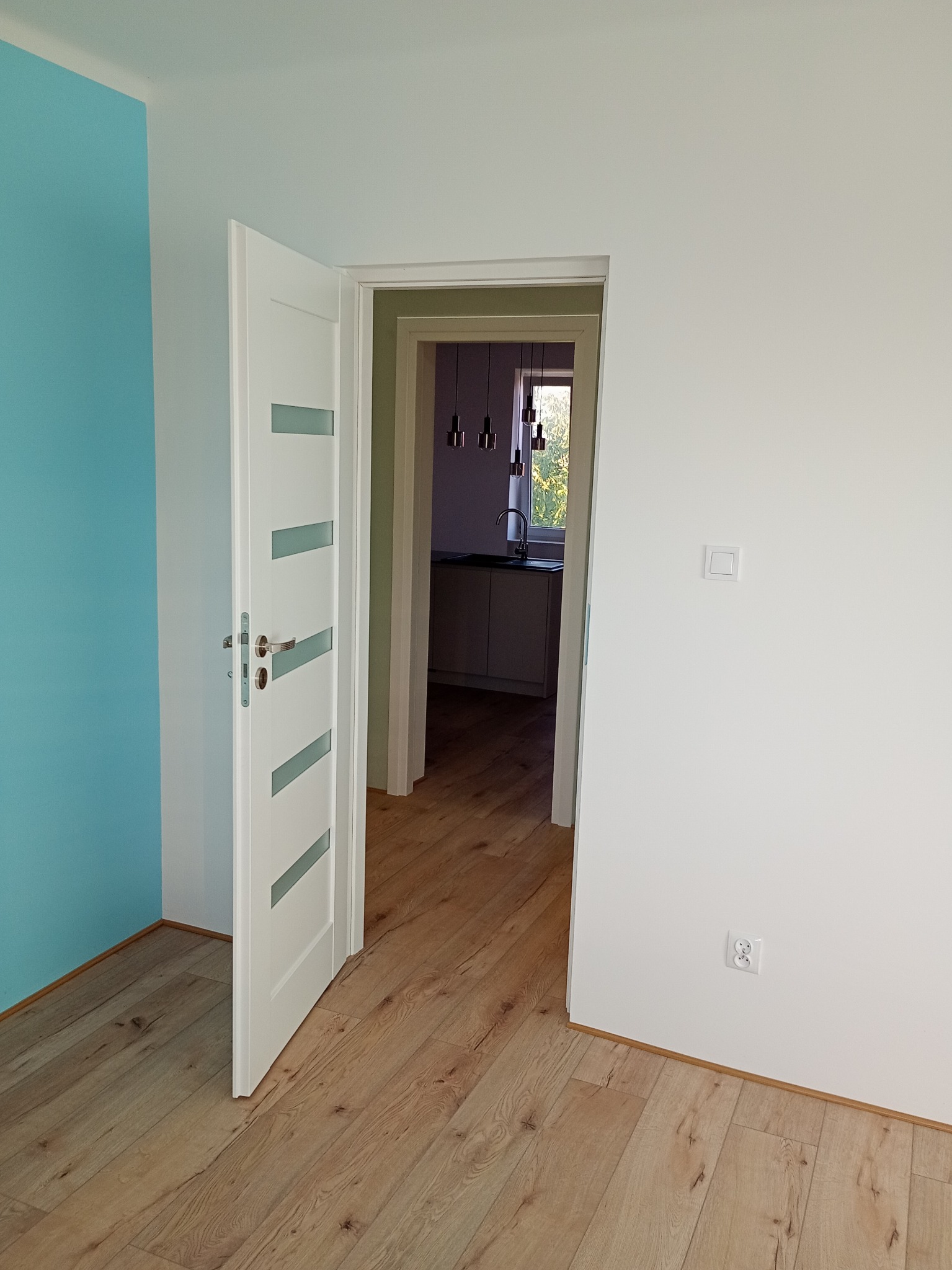 Jasne wnętrze po remoncie: świeżo pomalowane ściany w błękicie i bieli, drewniana podłoga, białe drzwi z przeszkleniami, widok na kuchnię z oknem.