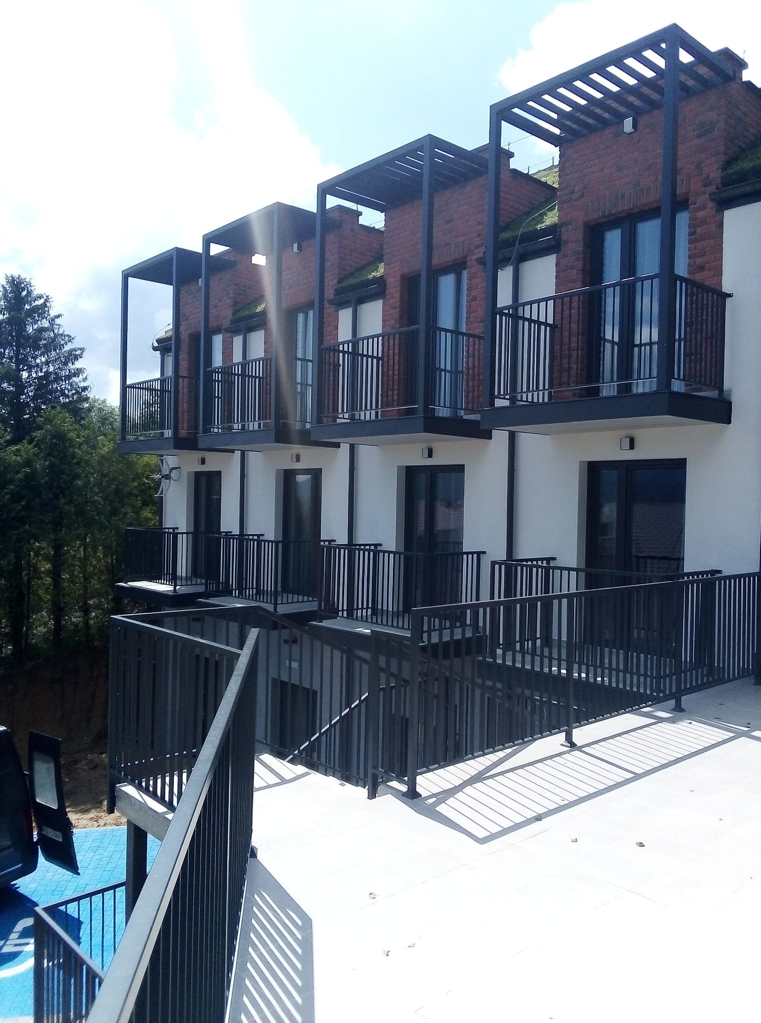 Nowoczesny budynek wielorodzinny z balkonami i elementami z cegły, widziany z dołu pod kątem. Metalowe balustrady i zadaszenia balkonów. Jasna elewacja i zieleń w tle.