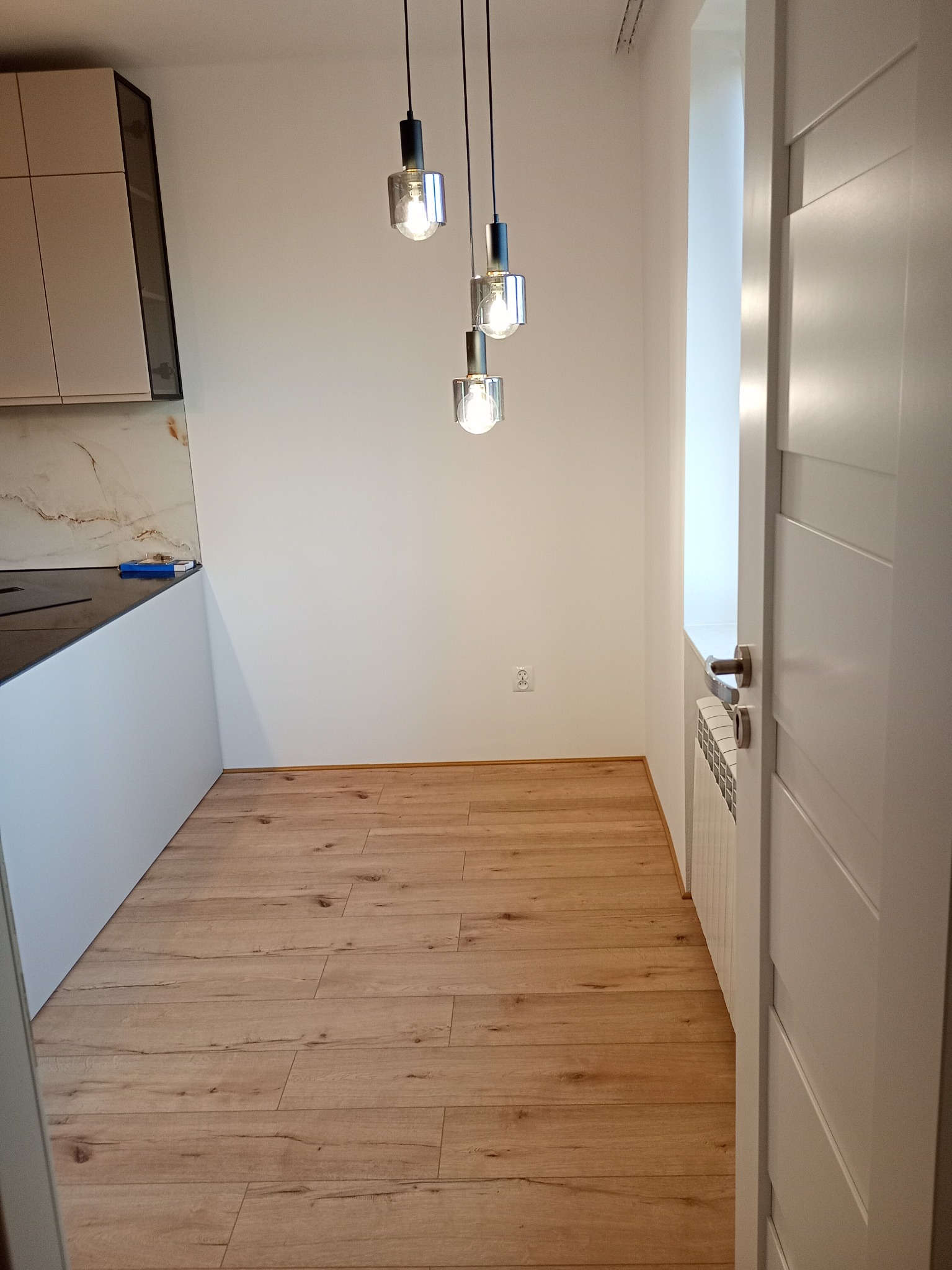 Jasne panele podłogowe w nowocześnie urządzonej kuchni z białymi ścianami, wiszącymi lampami i fragmentem zabudowy kuchennej. Widok na grzejnik i białe drzwi.