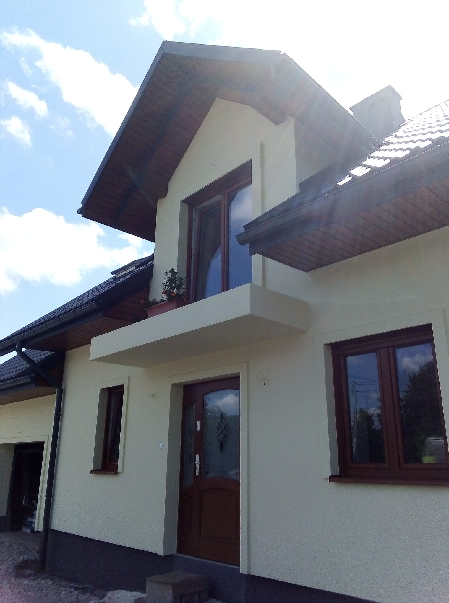 Elewacja domu jednorodzinnego po remoncie, z widocznymi oknami z brązowymi ramami, balkonem i świeżo pomalowaną fasadą w jasnym odcieniu beżu.