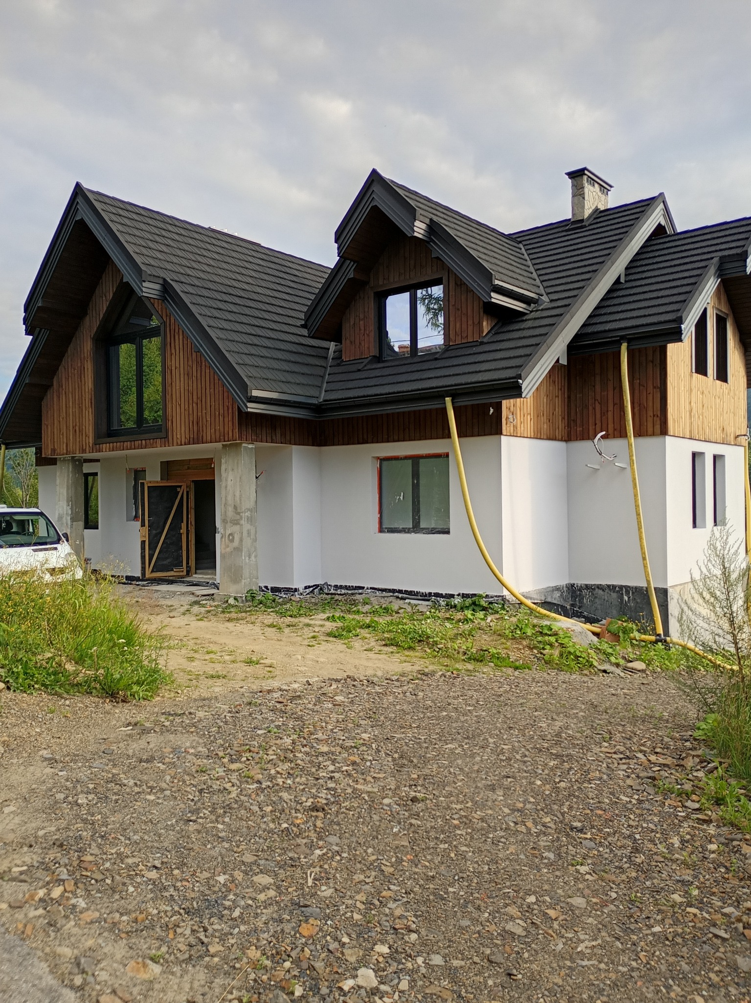 Nowoczesny dom z drewnianą elewacją i białym tynkiem, w trakcie wykańczania. Widoczne żółte węże i częściowo zagospodarowany teren wokół budynku.