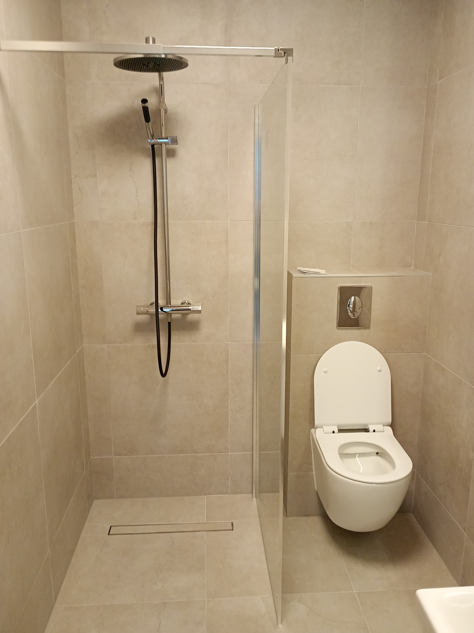 Nowoczesna łazienka w beżowych płytkach z kabiną prysznicową typu walk-in, podwieszaną toaletą i chromowaną armaturą. Minimalistyczny design, stonowana kolorystyka.