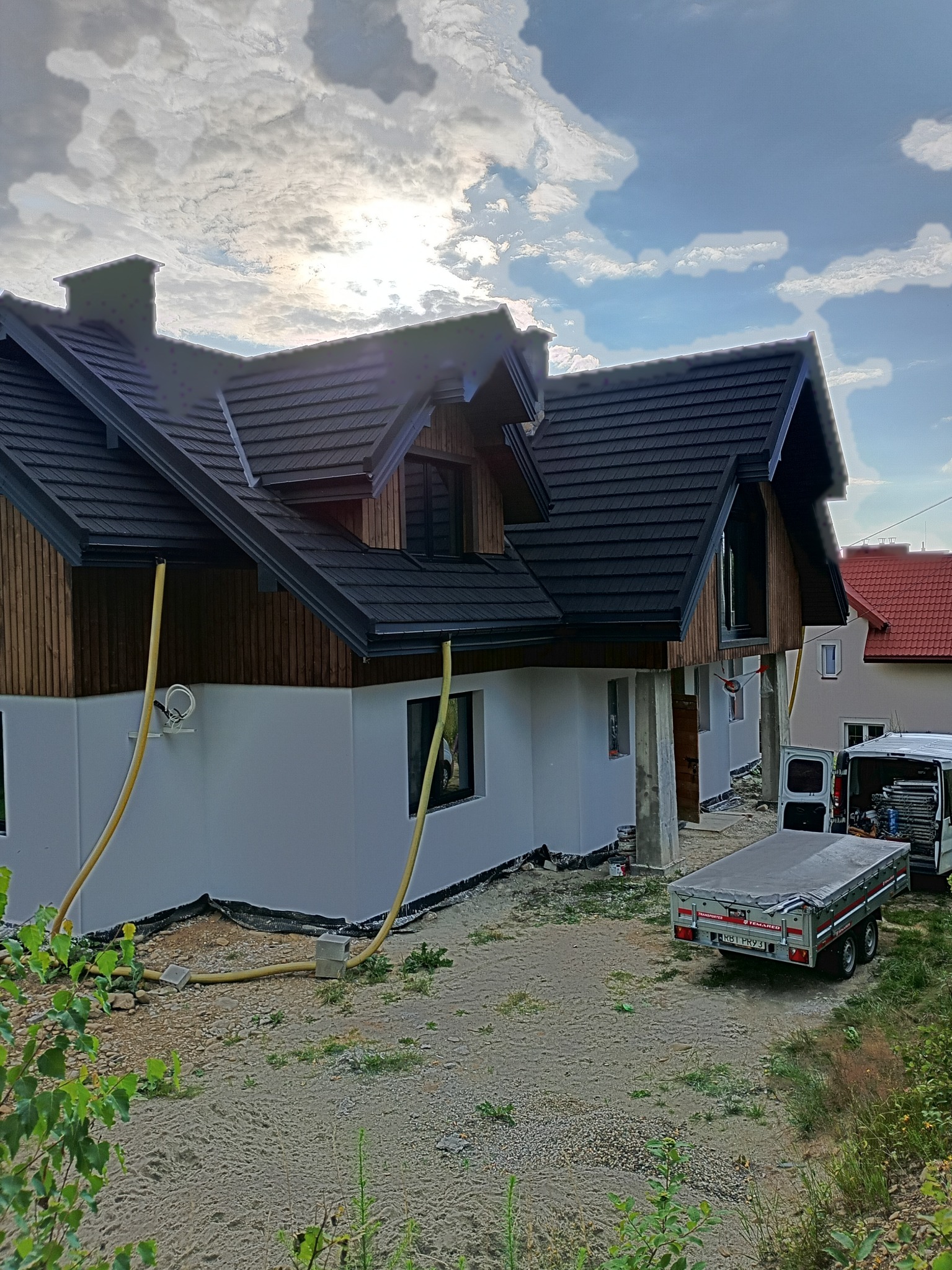 Dom po ociepleniu elewacji, widoczne rury do natrysku izolacji, przyczepka i bus w tle. Częściowo otynkowana elewacja, drewniana górna część budynku.