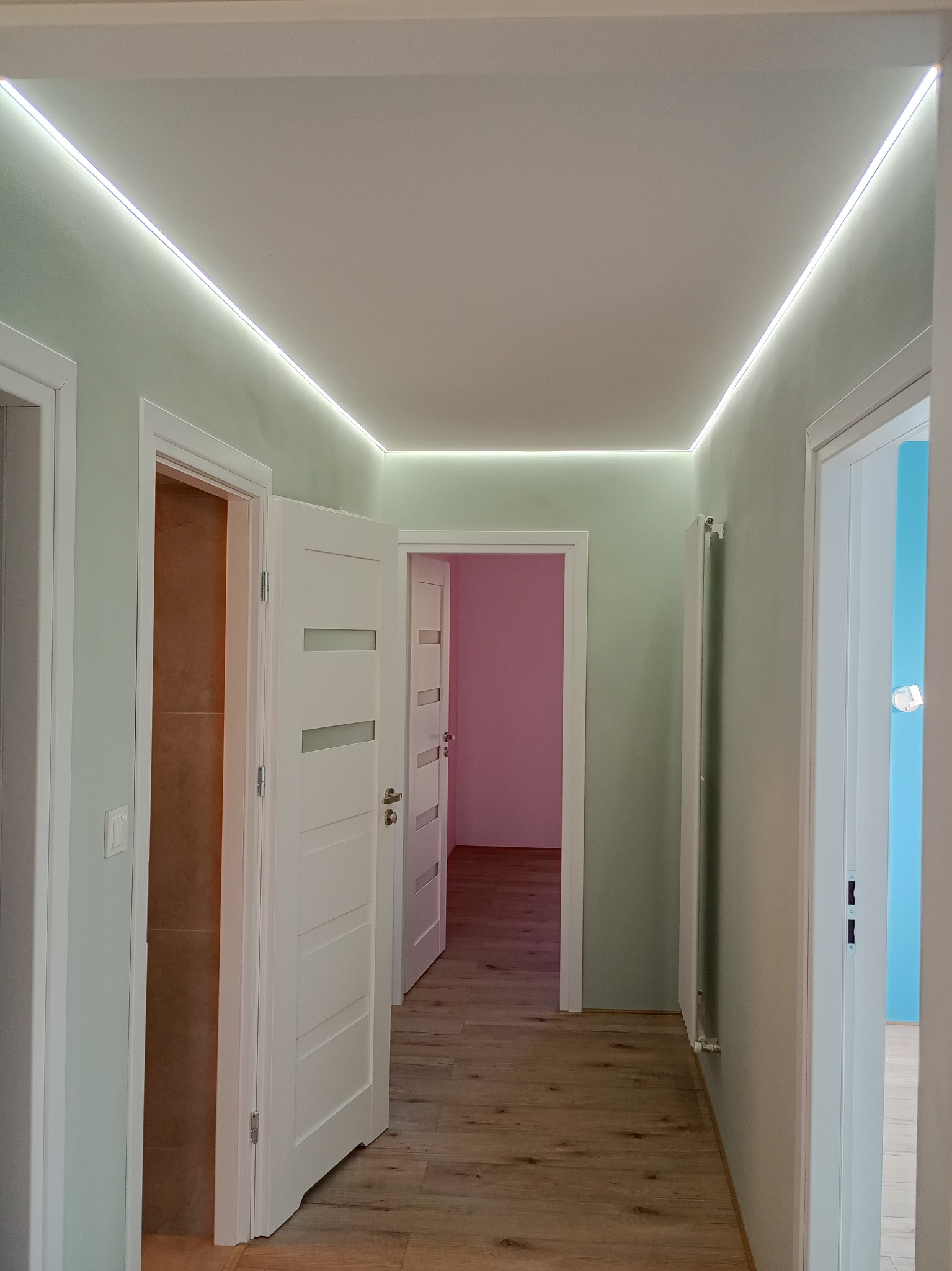 Korytarz po remoncie z oświetleniem LED na suficie, widoczne trzy białe drzwi do różnych pomieszczeń, podłoga z paneli. Ściany pomalowane na jasnozielony kolor.