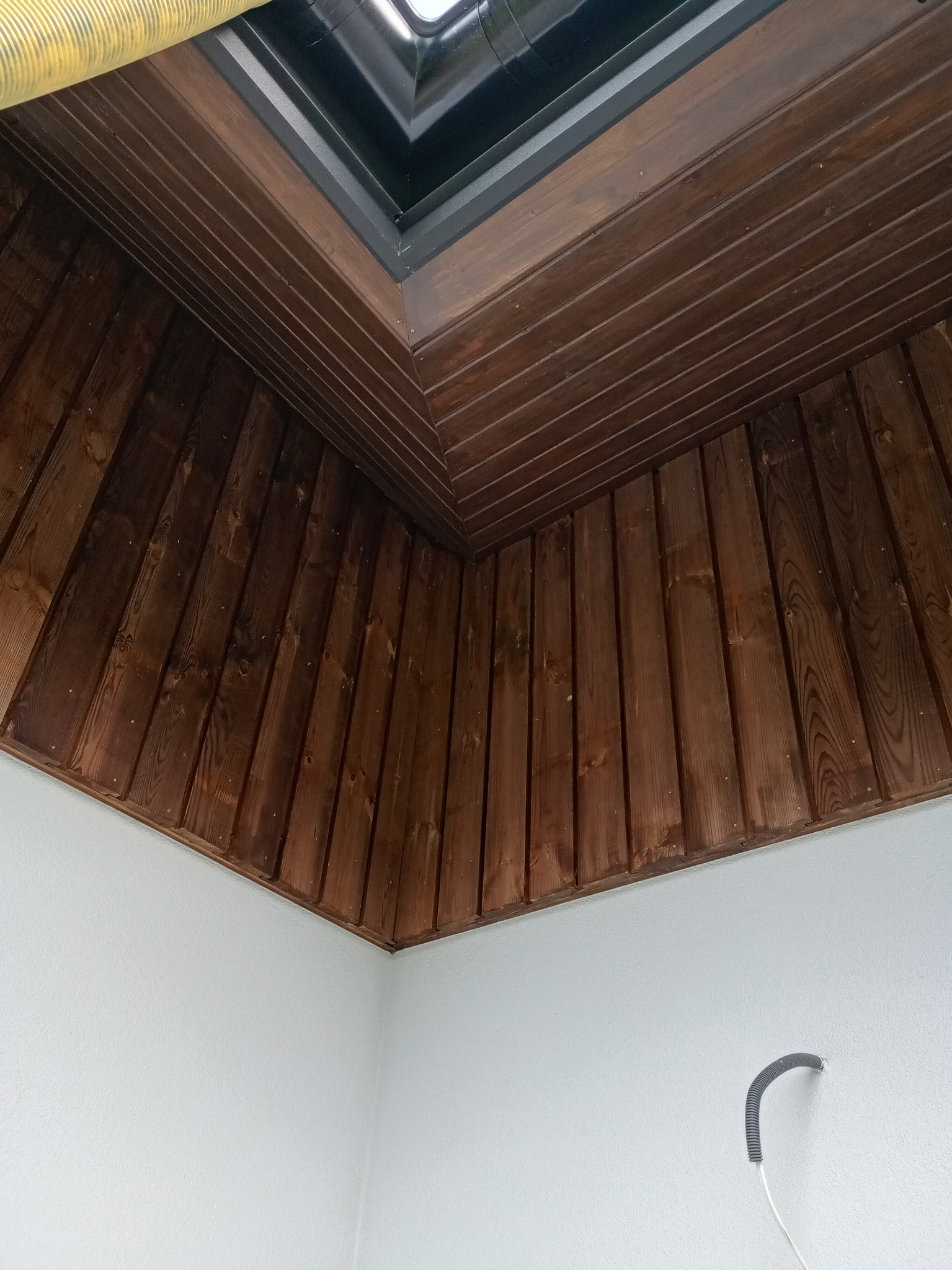 Wykończone drewnianą boazerią poddasze z oknem dachowym. Widoczne białe ściany i fragment instalacji. Ukośny sufit z ciemnego drewna.