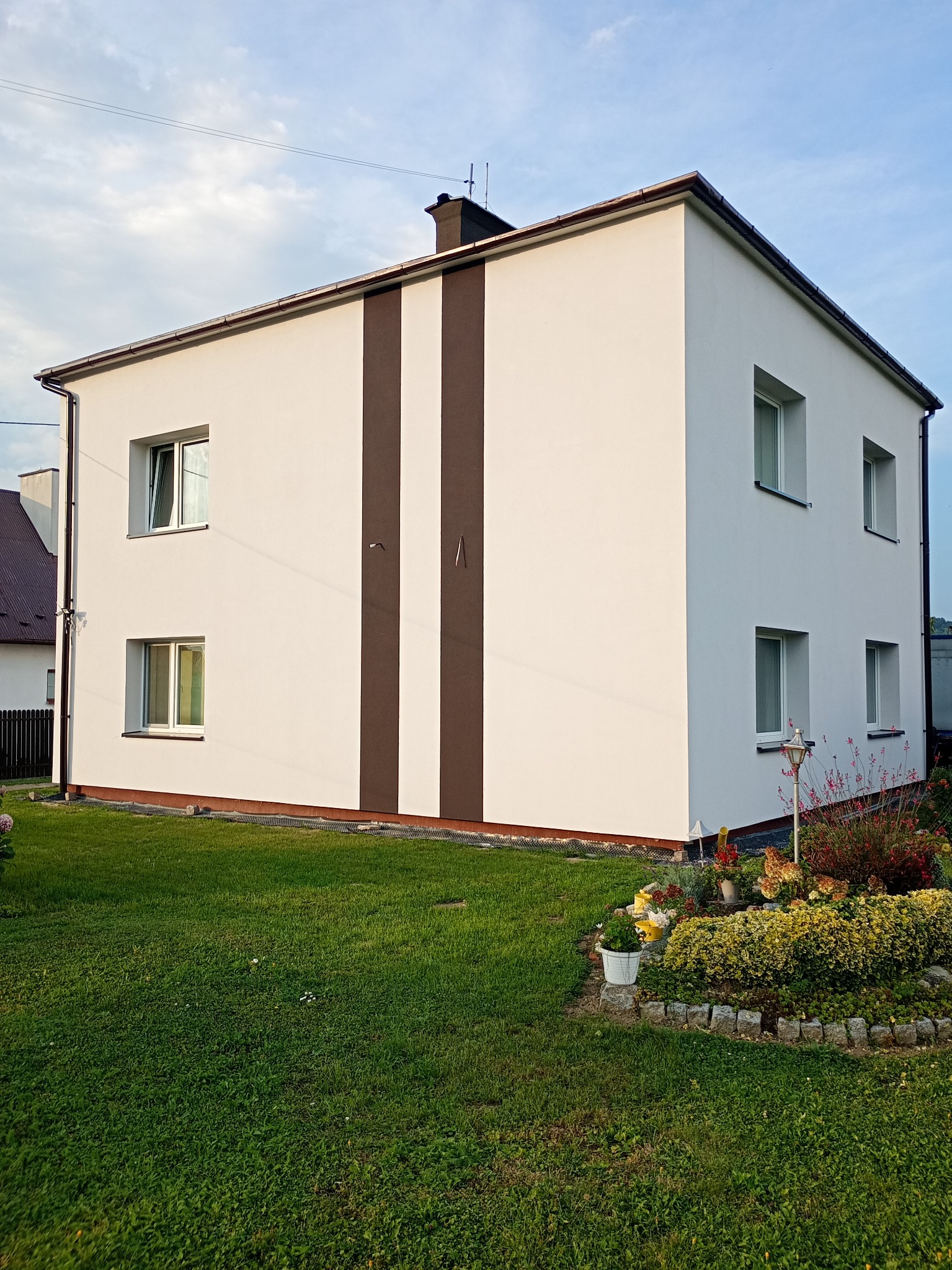 Dwukondygnacyjny dom z białą elewacją i brązowymi pasami, otoczony zielonym trawnikiem i rabatą kwiatową. Widoczne okna i elementy wykończenia dachu.