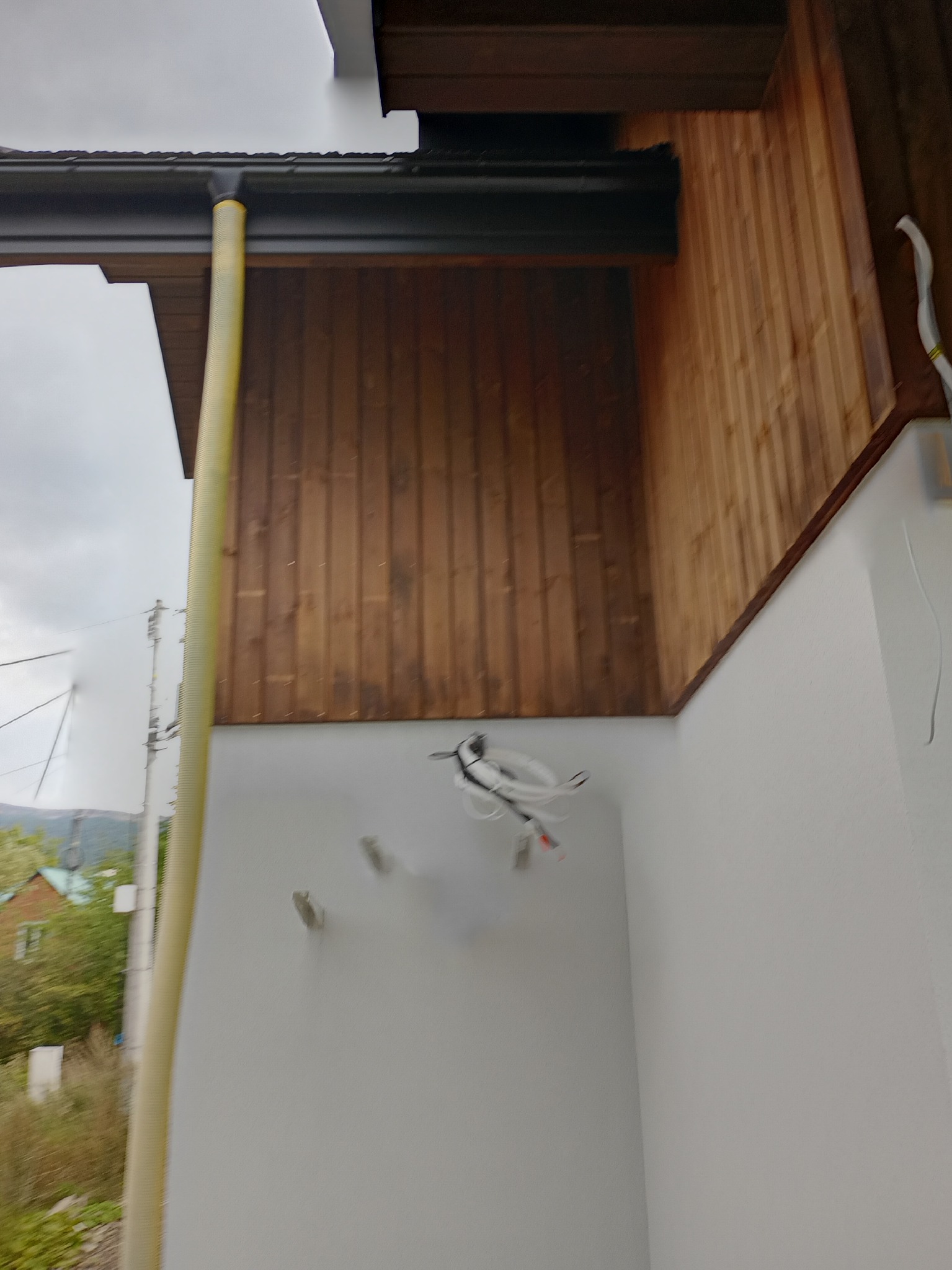 Fragment elewacji domu z drewnianą boazerią, rynną i wystającymi przewodami elektrycznymi. Widok z dołu, pochmurne niebo w tle.