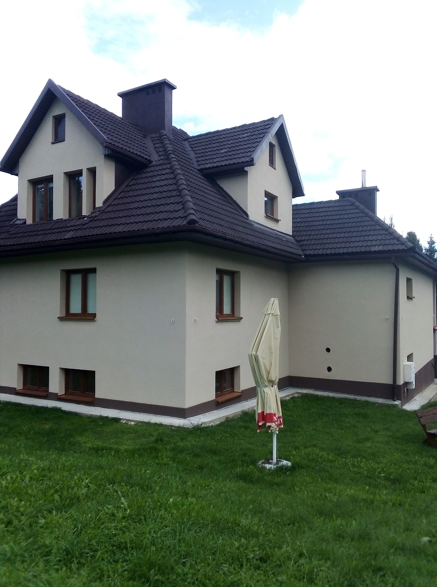 Dwukondygnacyjny dom z beżową elewacją i ciemnobrązowym dachem z dachówki, widoczny z poziomu trawnika. Zamknięty parasol w tle.