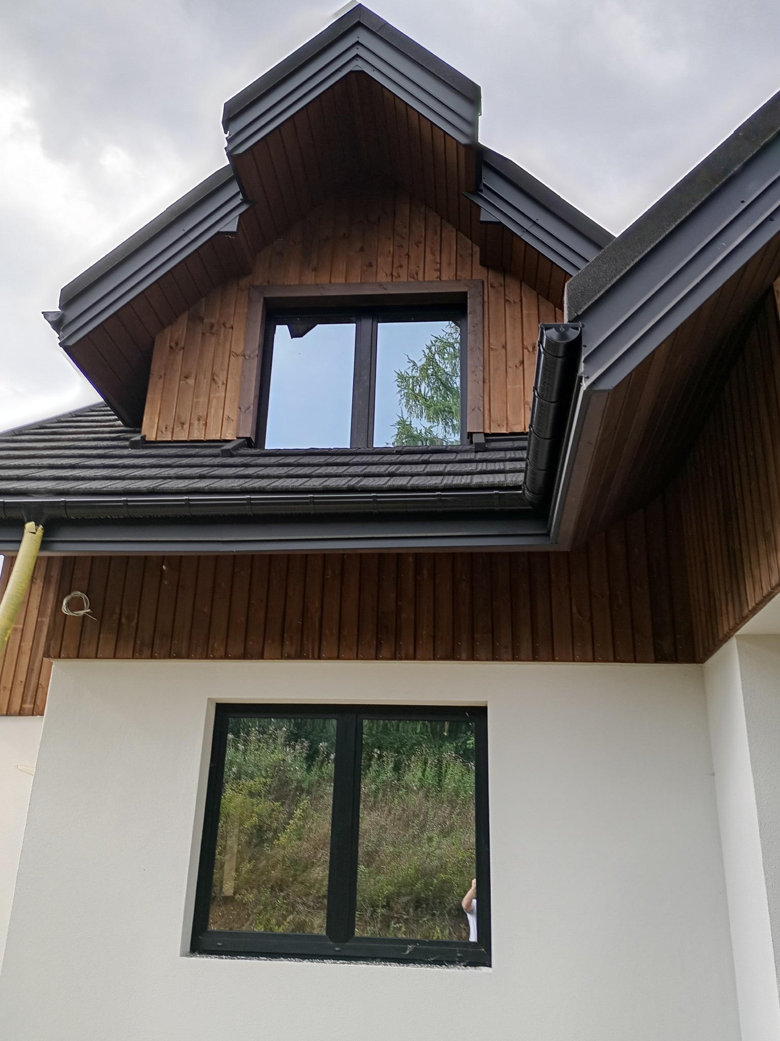 Wykończona elewacja domu z drewnianą okładziną i czarnymi ramami okiennymi. Widoczne orynnowanie i fragment dachu. Biała ściana na dole kadru.