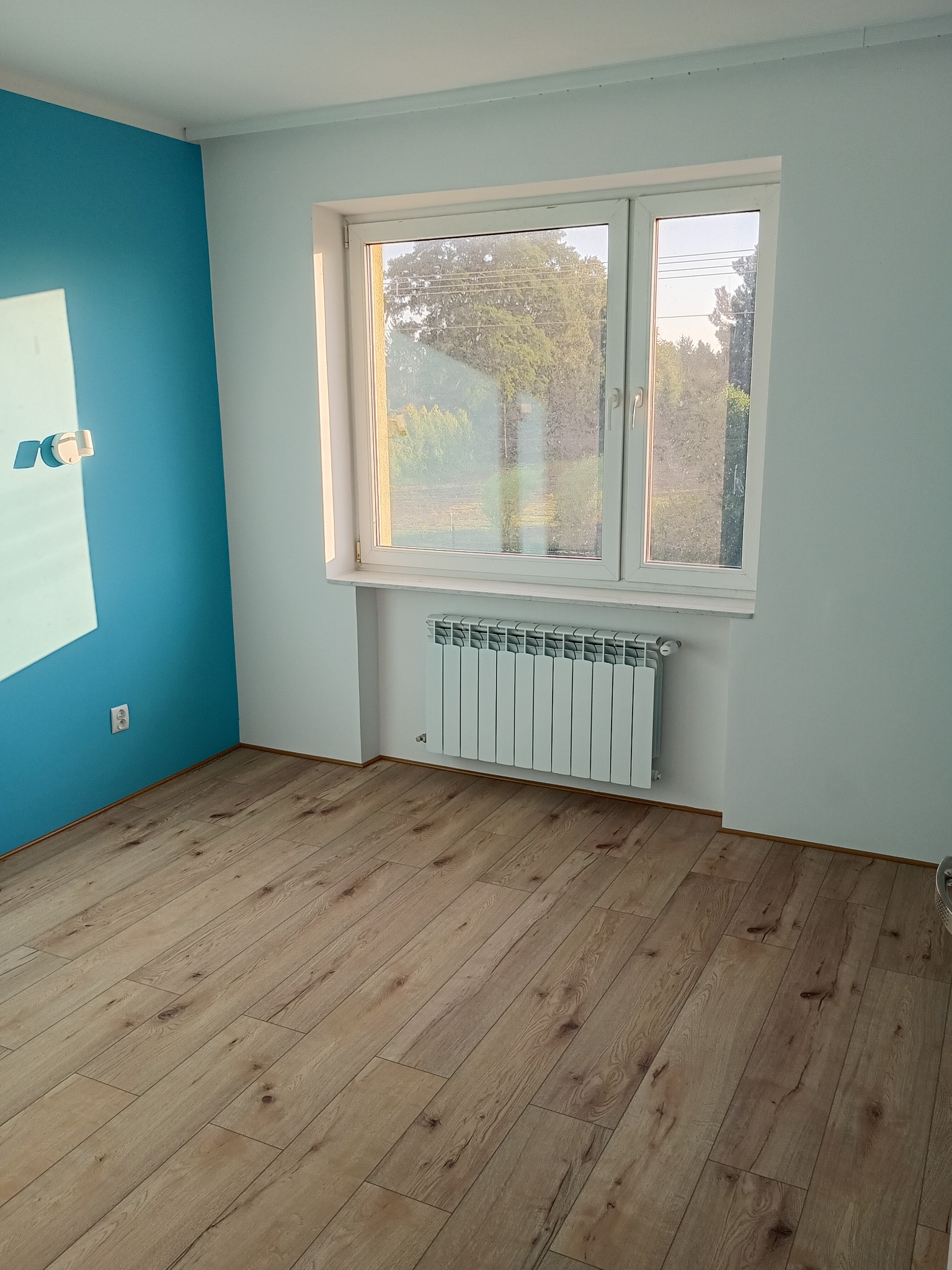 Pomalowany pokój z panelami podłogowymi i grzejnikiem pod oknem. Jedna ściana w kolorze turkusowym, pozostałe białe. Widoczne gniazdko elektryczne i włącznik światła.
