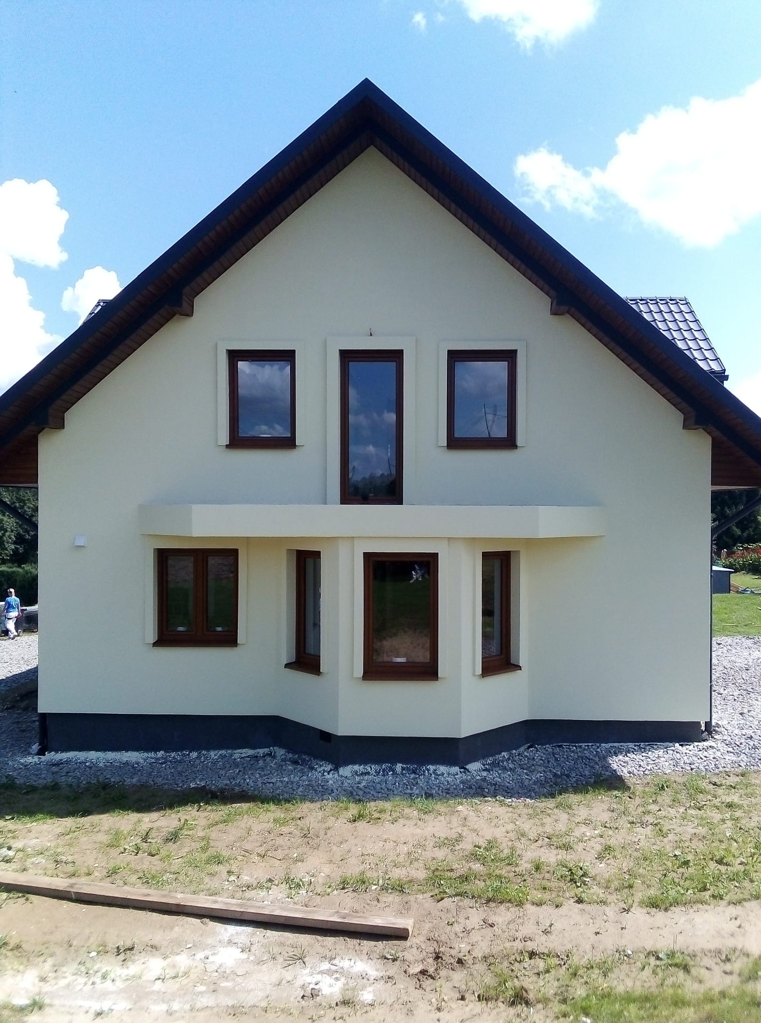 Elewacja domu jednorodzinnego po ociepleniu, z widocznymi brązowymi ramami okiennymi i ciemnym dachem. Jasna fasada, błękitne niebo w tle.