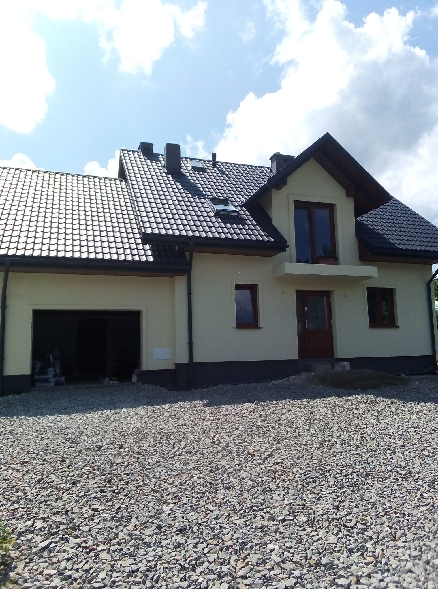 Jasny dom jednorodzinny z ciemnym dachem i otwartym garażem, otoczony żwirowym podłożem. Widoczne okna, drzwi wejściowe i fragment nieba z chmurami.