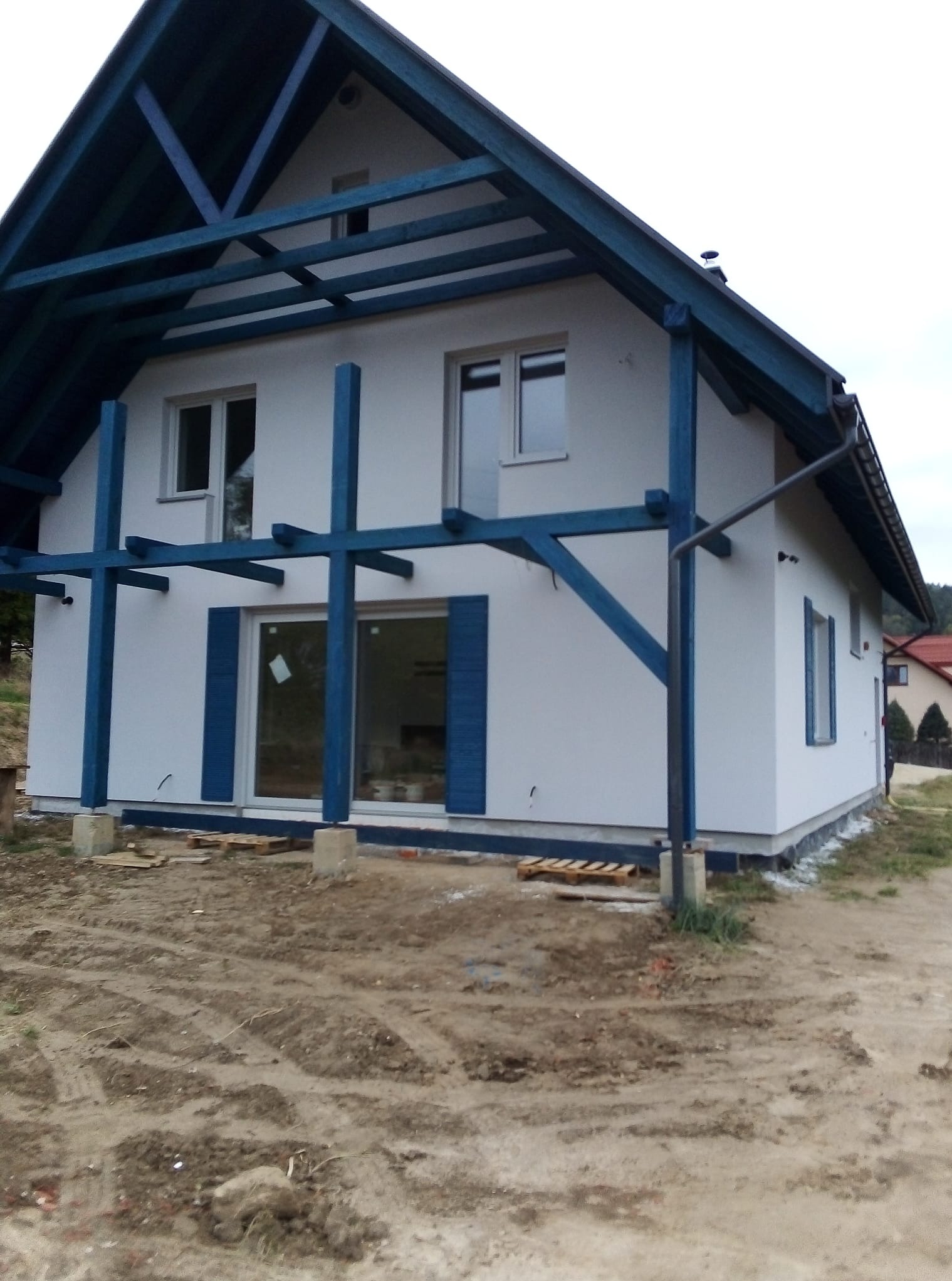 Biały dom z niebieskimi elementami konstrukcyjnymi i okiennicami, widok z zewnątrz na tle ziemi. Dom w trakcie wykończenia, widoczne podpory i palety.