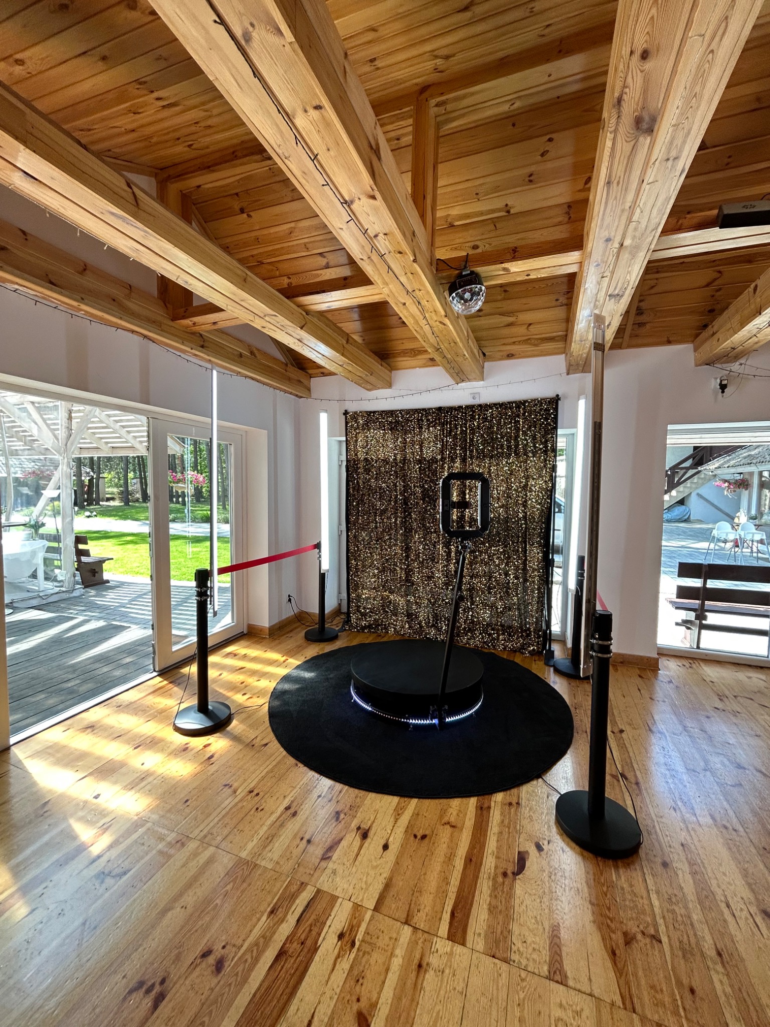 Wnętrze sali z drewnianym sufitem i podłogą, przygotowane do imprezy z instalacją do robienia zdjęć na czarnym podeście, z błyszczącą ścianką w tle i kulą dyskotekową pod sufitem.