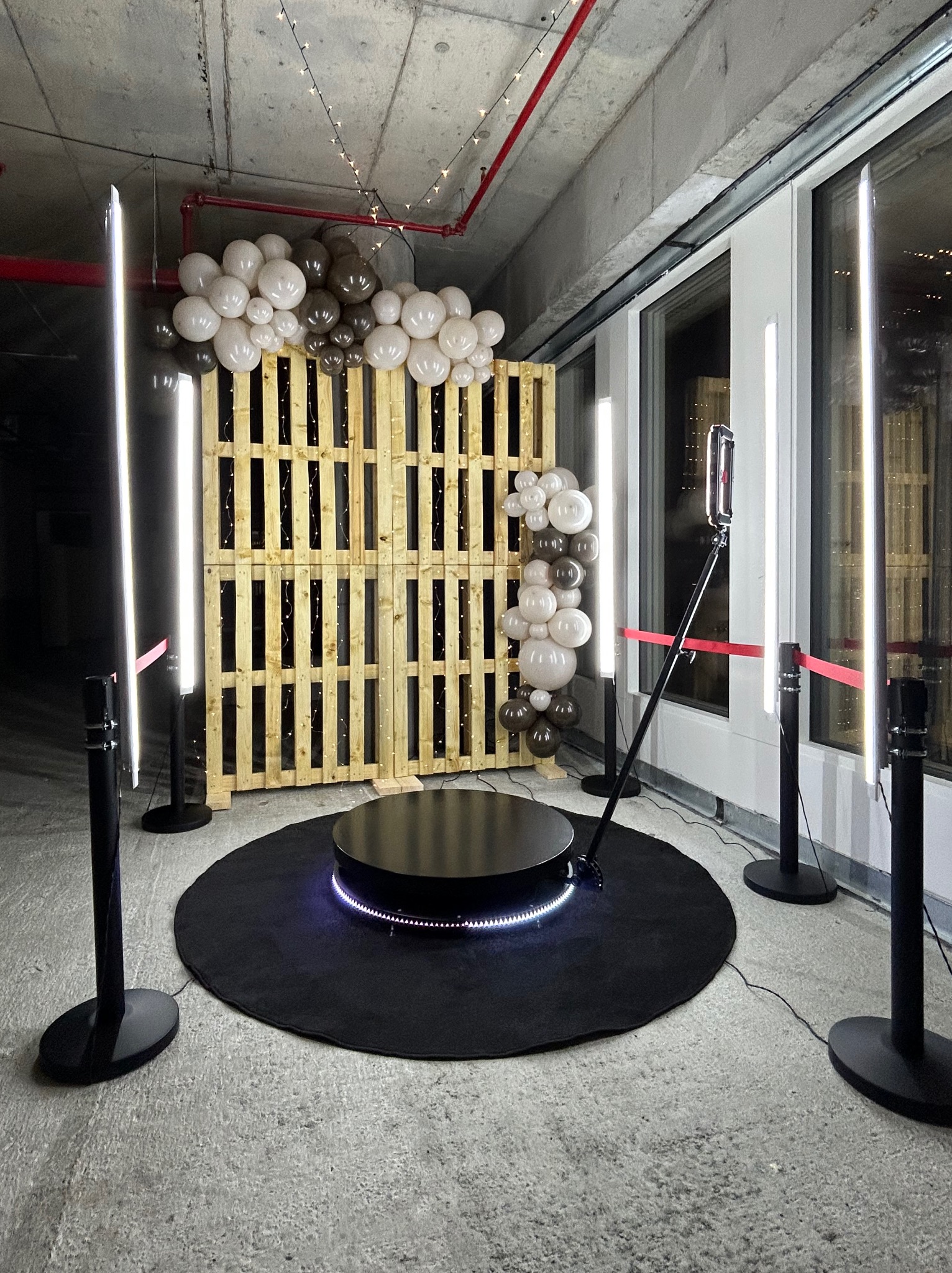Instalacja z obrotową platformą na czarnym dywanie, drewniana paleta z lampkami i balonami w odcieniach bieli i brązu, oświetlenie ledowe, automat do zdjęć na statywie.