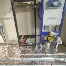 Marcin Szczęch - Montaż stelaża podtynkowego Geberit z instalacją ogrzewania podłogowego, widoczne rury doprowadzające wodę, folia aluminiowa pod wężami grzewczymi.