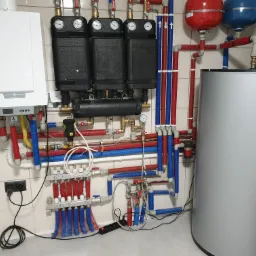 Kompleksowa instalacja grzewcza z kotłem gazowym, rozdzielaczami, zbiornikiem na wodę i licznymi rurami w kolorach czerwonym i niebieskim, zamontowana na kafelkowanej ścianie.