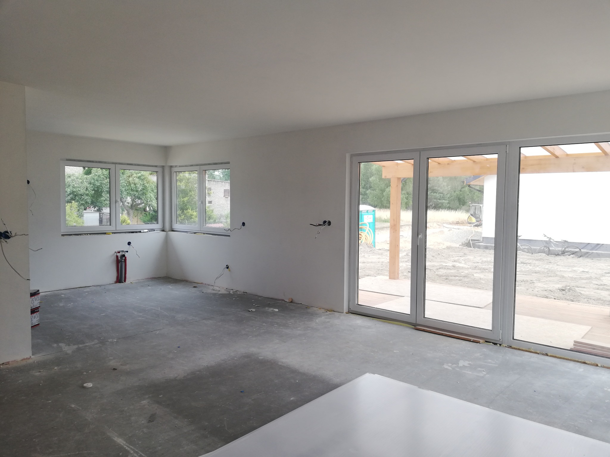 Surowe wnętrze domu w trakcie wykończenia: gładkie, białe ściany, betonowa podłoga, duże okna z widokiem na zewnątrz i wystające kable elektryczne.