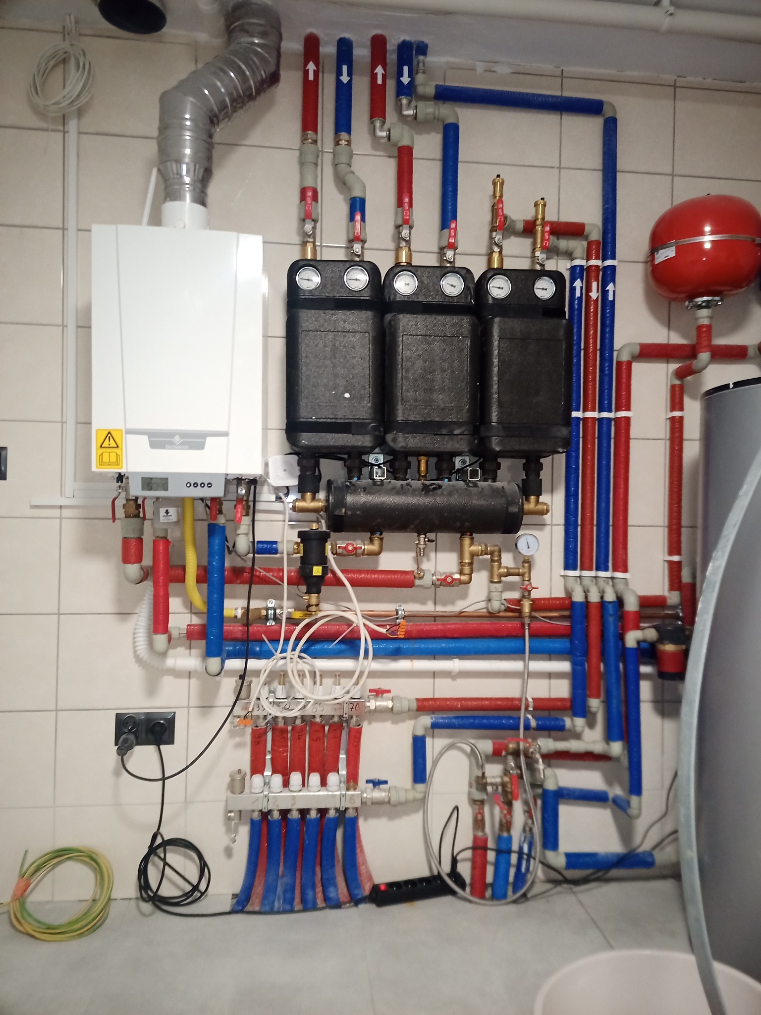 Kompletna instalacja centralnego ogrzewania z kotłem gazowym, rozdzielaczem, pompami i zbiornikiem wyrównawczym, z rurami oznaczonymi kolorami czerwonym i niebieskim, zamontowana na ścianie...