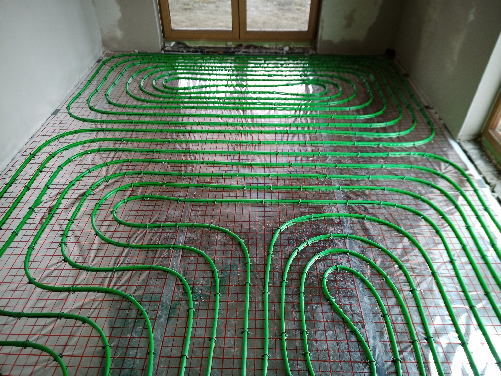 Zielone rury ogrzewania podłogowego ułożone na folii z czerwoną siatką, widoczne okno i surowa ściana w tle.