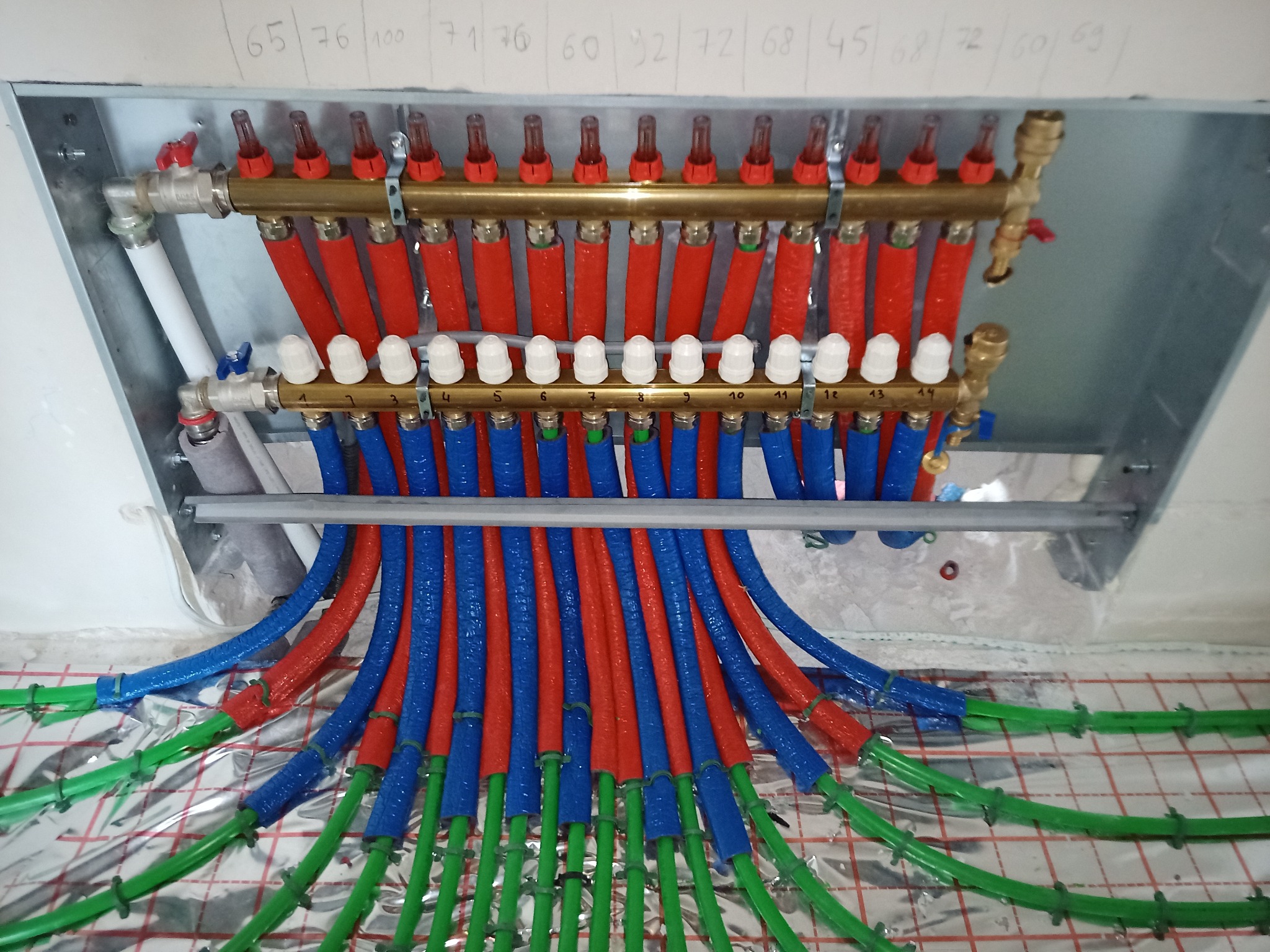 Rozdzielacz ogrzewania podłogowego z rurami w kolorach czerwonym, niebieskim i zielonym, zamontowany na folii izolacyjnej z siatką, widoczne numery na rozdzielaczu i zapisane wartości na ścianie.