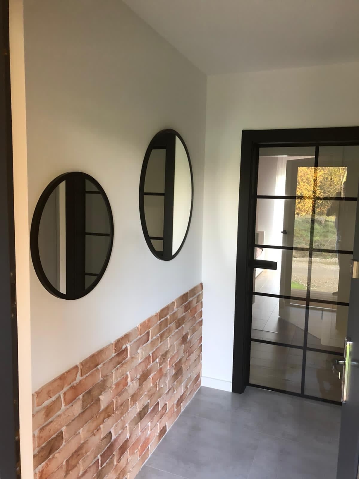 Korytarz z dekoracyjną ścianą z cegły, dwoma okrągłymi lustrami z czarnymi ramami oraz czarnymi drzwiami z przeszkleniami w stylu industrialnym.