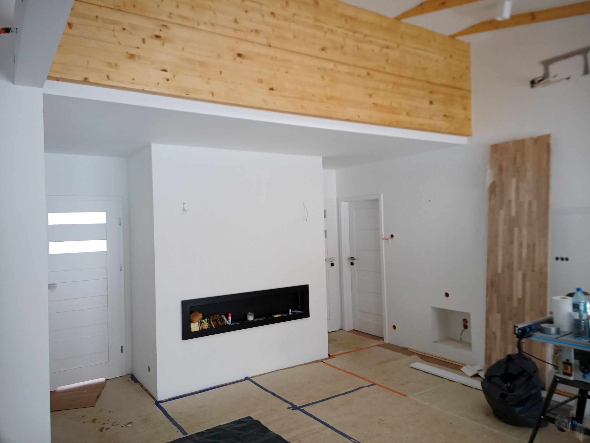Wnętrze w trakcie wykańczania: widoczna drewniana konstrukcja sufitu, ściany pomalowane na biało, wnęka na kominek, częściowo wykończona ściana z drewnianych paneli, przygotowane podłoże pod posadzkę.